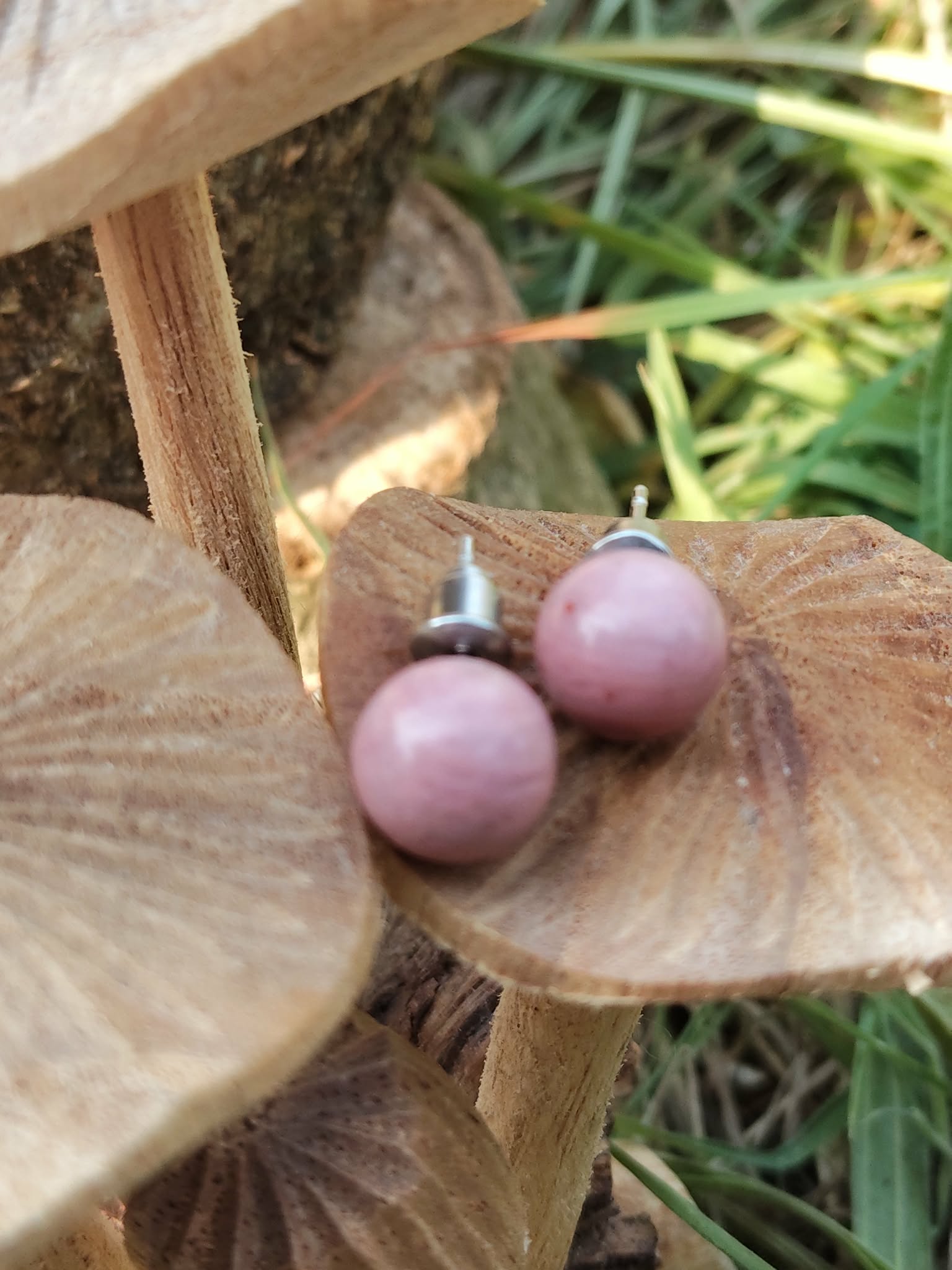 Boucles d’oreilles puces rhodonite rose, tiges acier inoxydable, posées sur support en bois avec feuillage en arrière-plan; douceur, apaisement émotionnel.