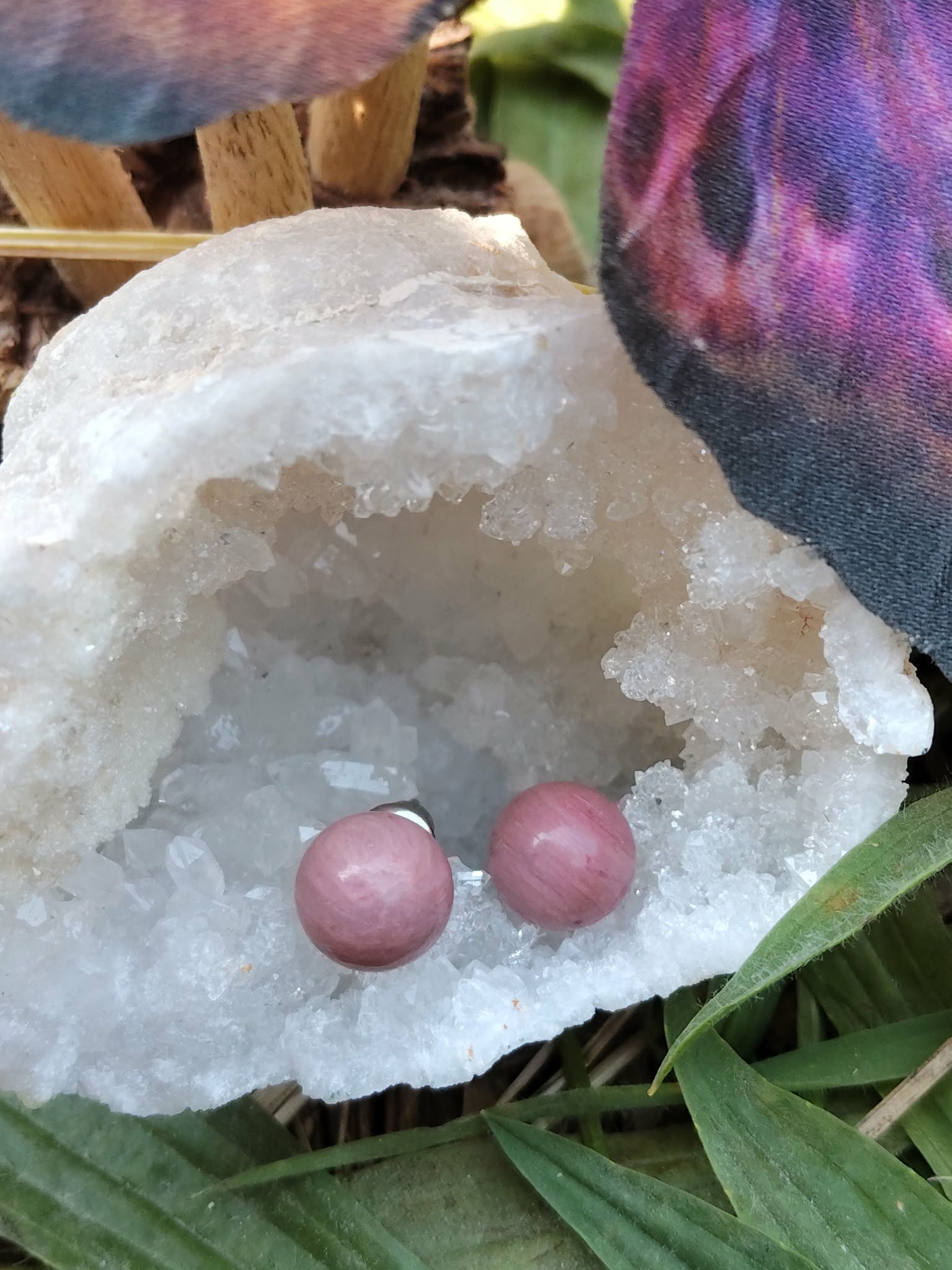 Boucles d’oreilles puces en rhodonite rose (perles rondes) sur tiges en acier inoxydable, posées au creux d’une géode de quartz avec feuillage en décor; ambiance douce, réconfortante, favorise l’apaisement émotionnel et la bienveillance.