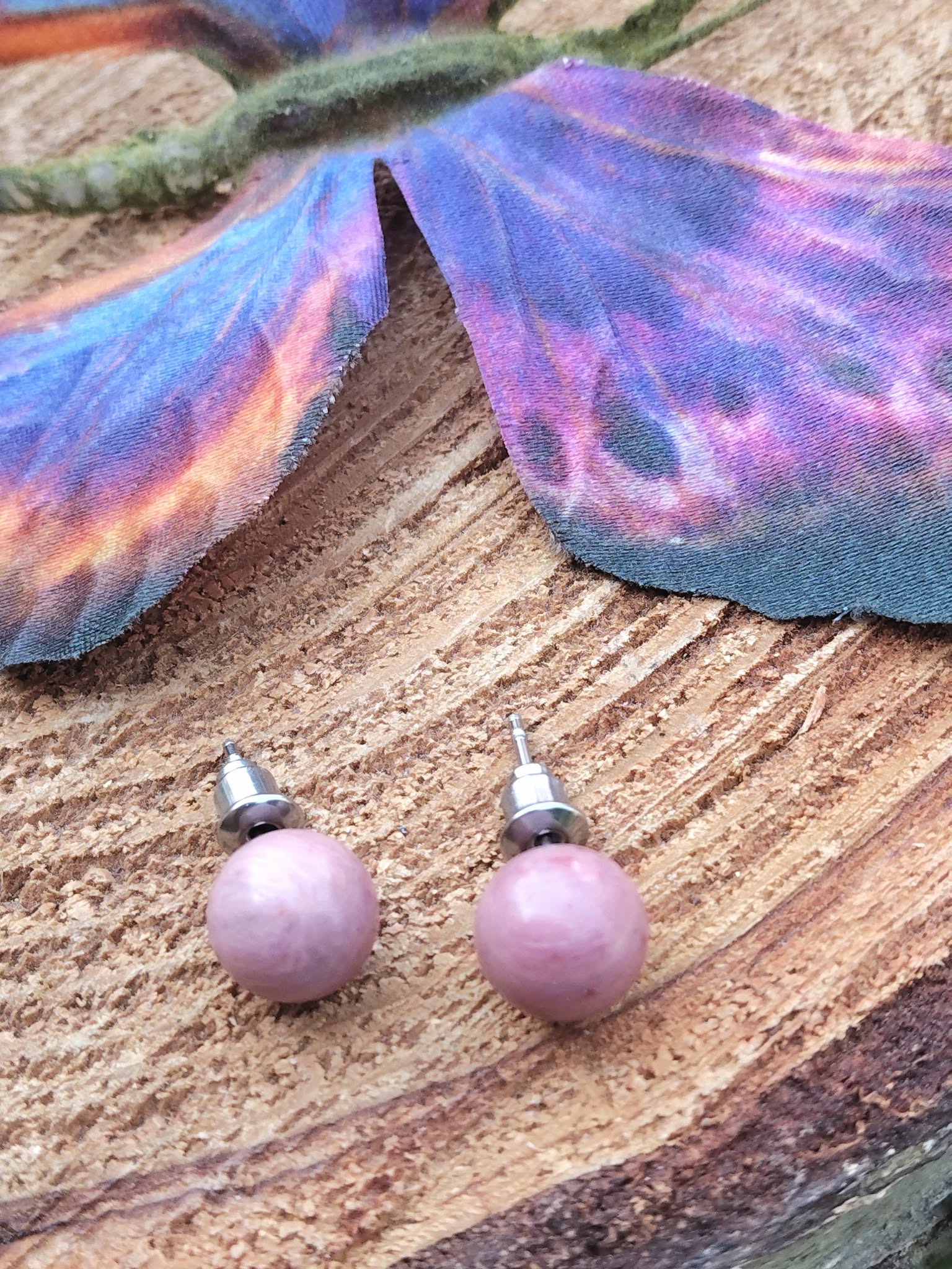 Boucles d’oreilles puces rhodonite rose, tiges acier inoxydable, sur tranche de bois avec papillon violet/bleu en arrière-plan; douceur et apaisement émotionnel.