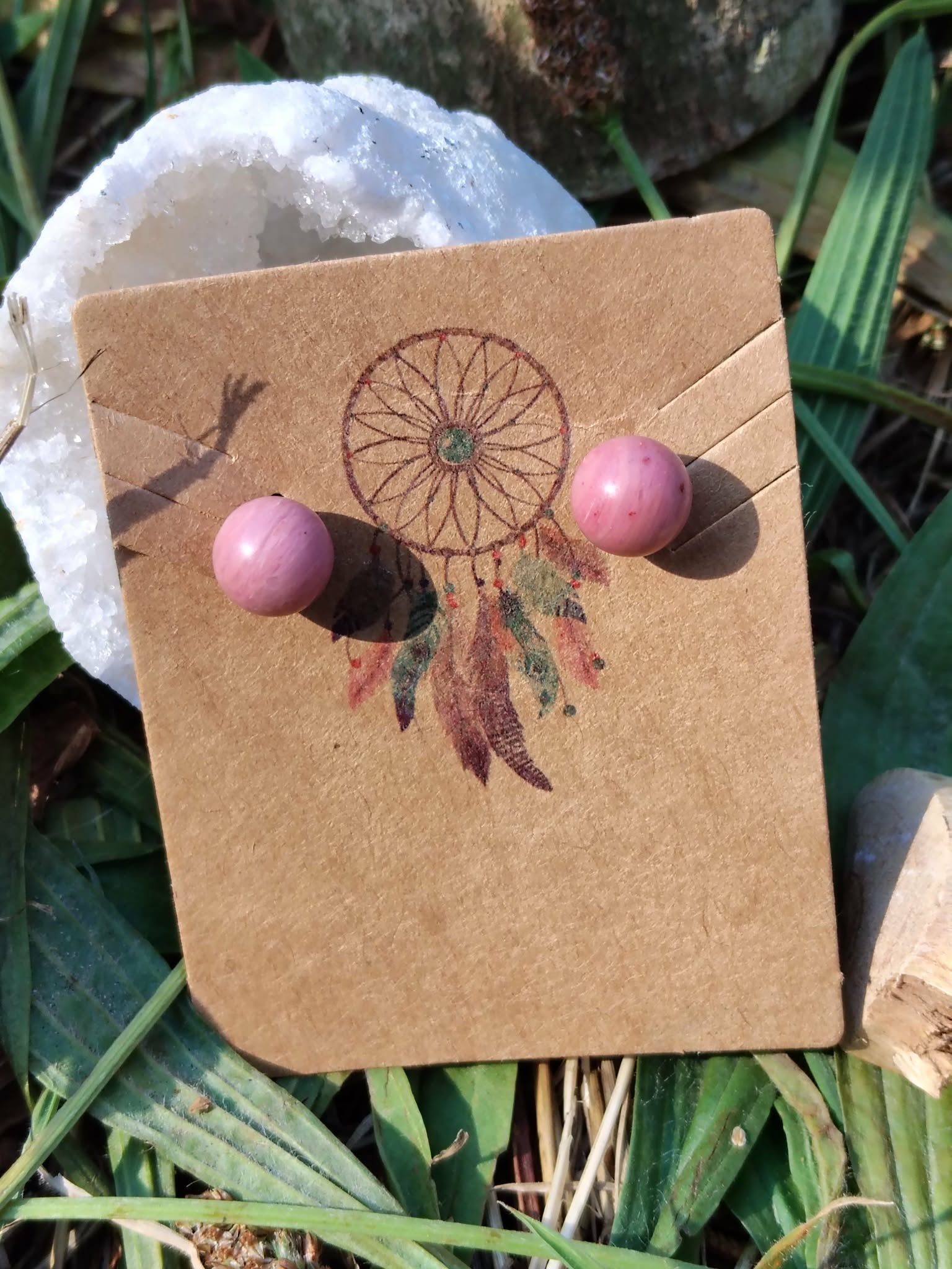 Boucles d’oreilles puces rhodonite rose, tiges acier inoxydable, sur carte kraft attrape-rêves avec géode et feuillage; évoque douceur et apaisement émotionnel.