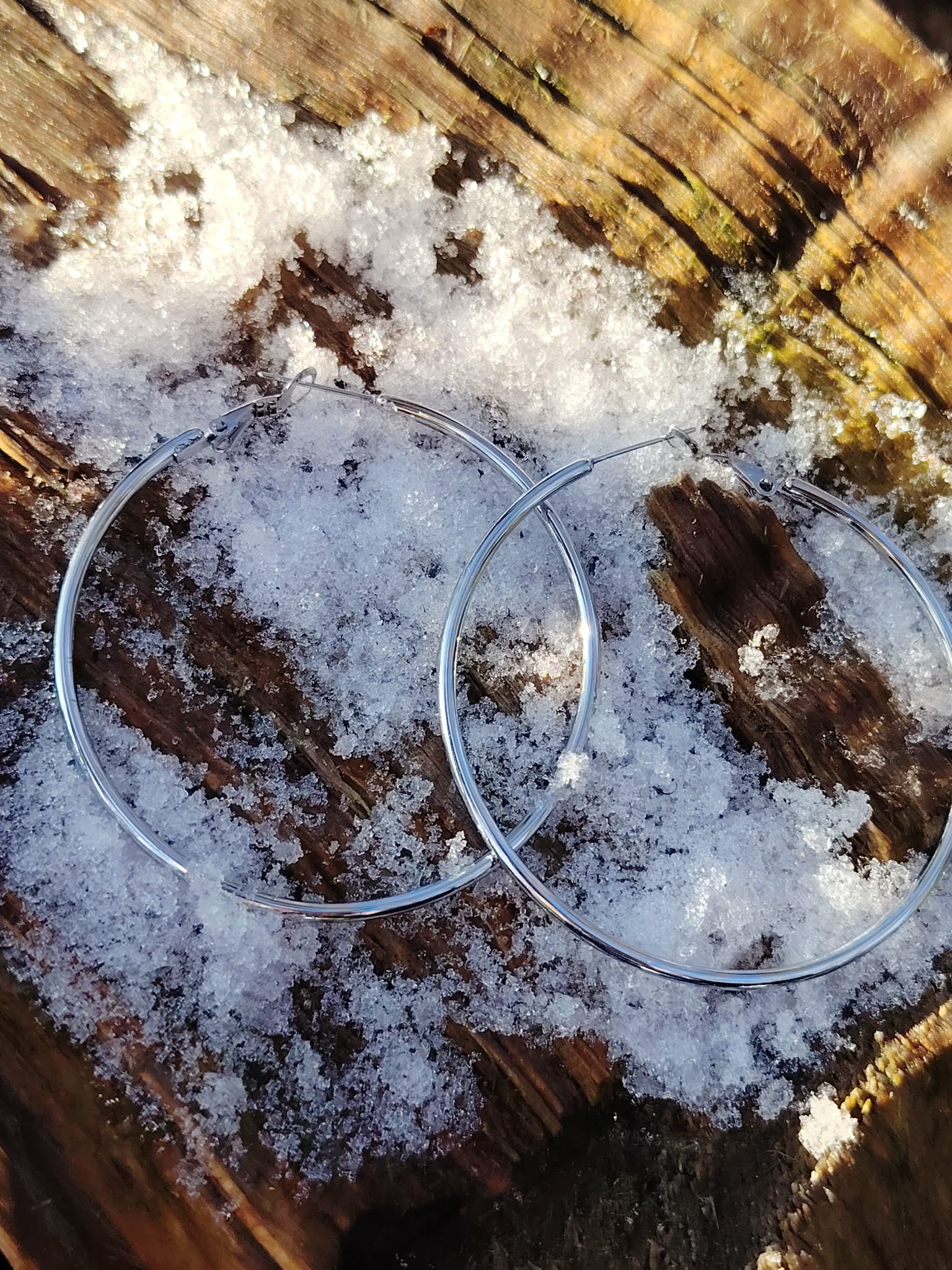 Boucles d’oreilles grandes créoles argentées en acier inoxydable, posées sur bois avec neige fondue; photo prise en Bretagne; évoquent fraîcheur, renouveau, clarté.