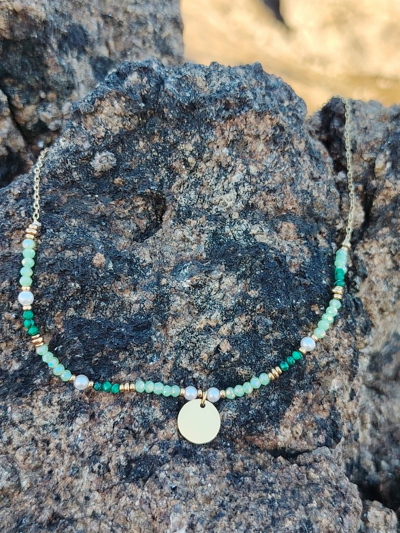 Collier doré à cristaux verts sur rochers sombres en bord de mer en Bretagne, équilibre visuel et allure naturelle raffinée