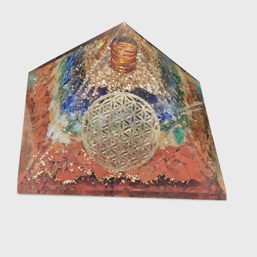 Pyramide orgonite 7 chakras avec fleur de vie et spirale de cuivre, inclusions multicolores et paillettes dorées, photo sur fond blanc ; évoque harmonie, ancrage, protection.