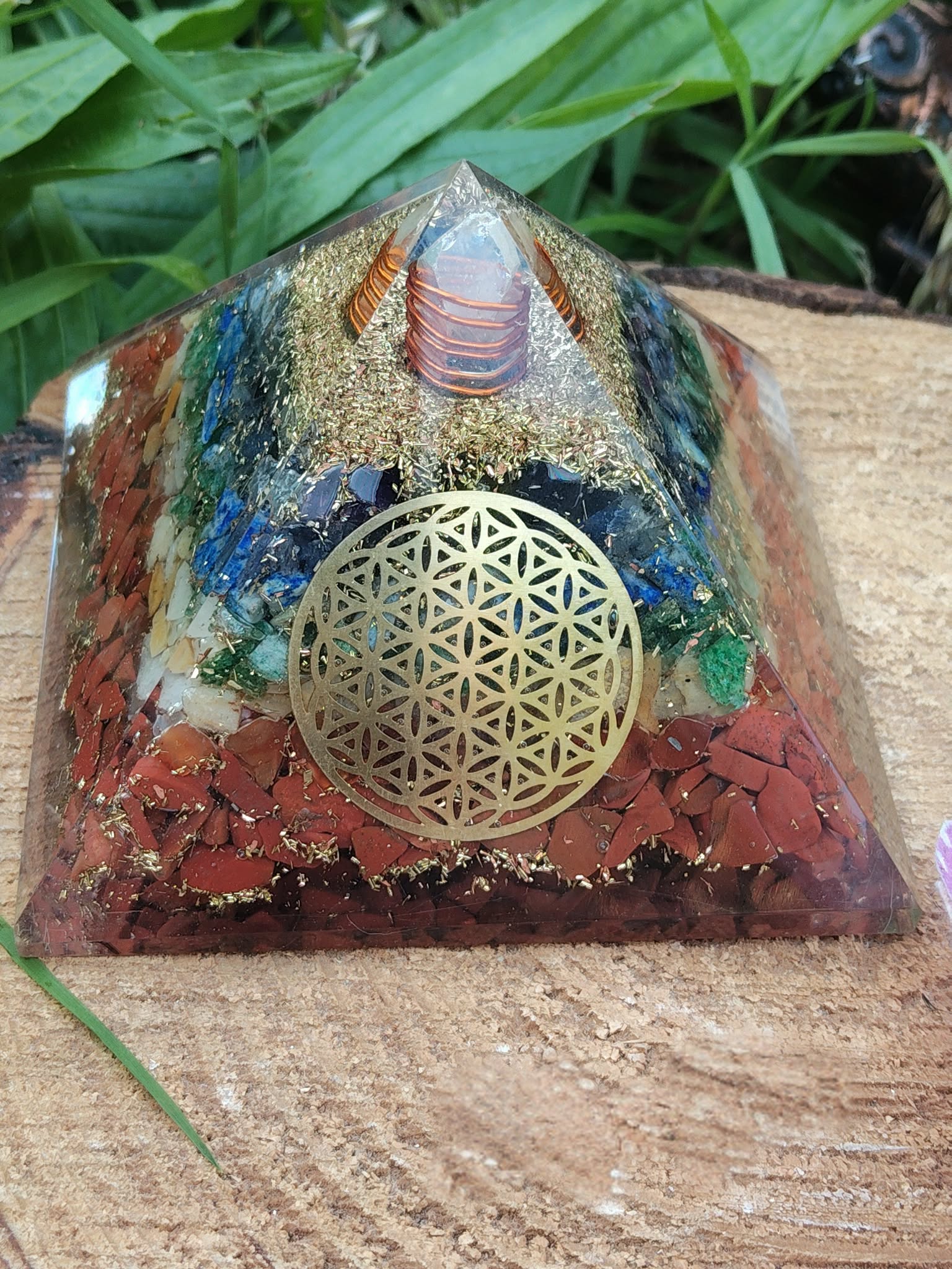 Pyramide orgonite 7 chakras avec fleur de vie et spirale de cuivre, posée sur une tranche de bois avec herbe en arrière-plan ; évoque harmonie, équilibre, ancrage.