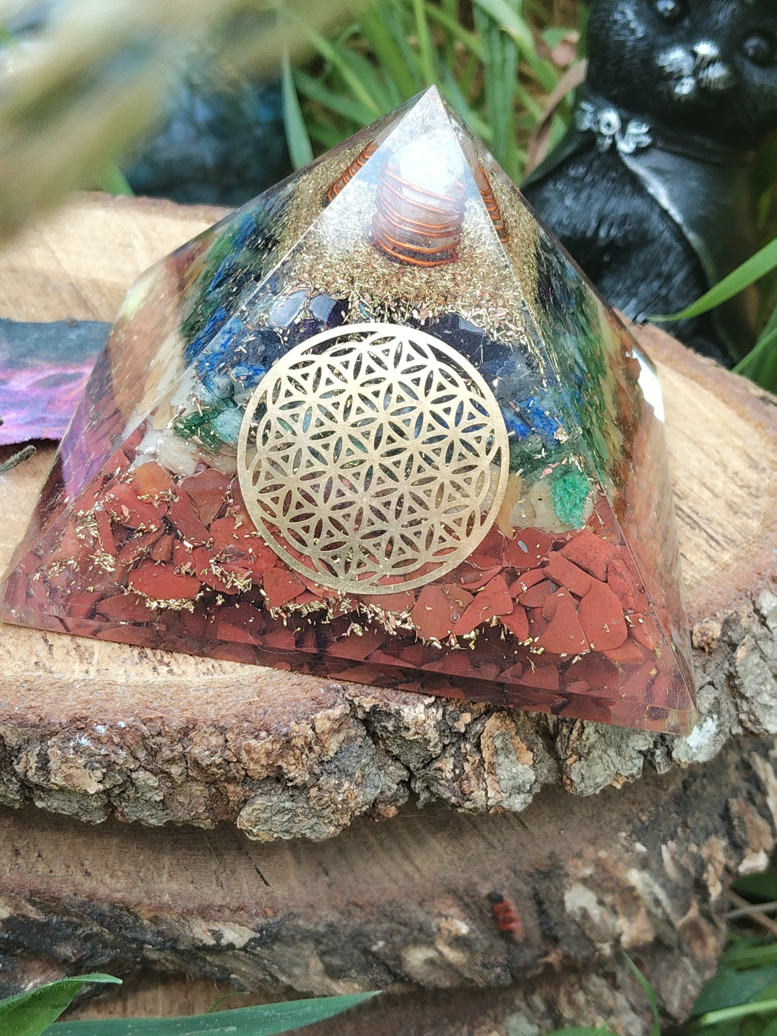 Pyramide orgonite 7 chakras avec fleur de vie et spirale de cuivre, posée sur une tranche de bois en extérieur (herbe, pierre au premier plan) ; évoque harmonie, ancrage, protection.