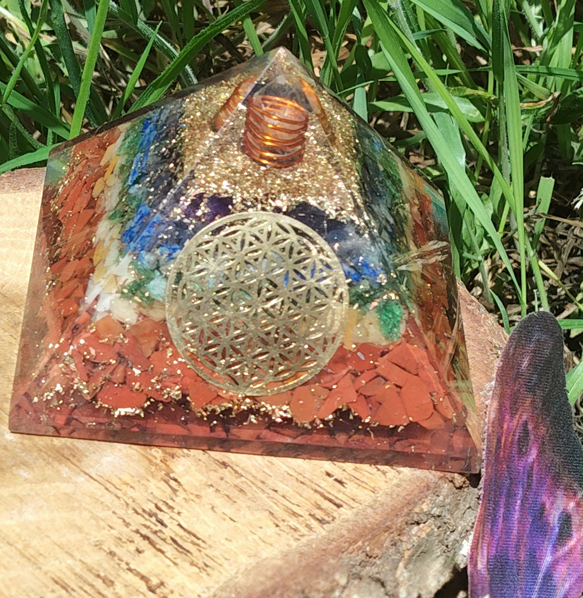 Pyramide orgonite 7 chakras avec fleur de vie et spirale de cuivre, posée sur tranche de bois en extérieur (herbe, pierre violette) ; évoque équilibre, ancrage, protection.