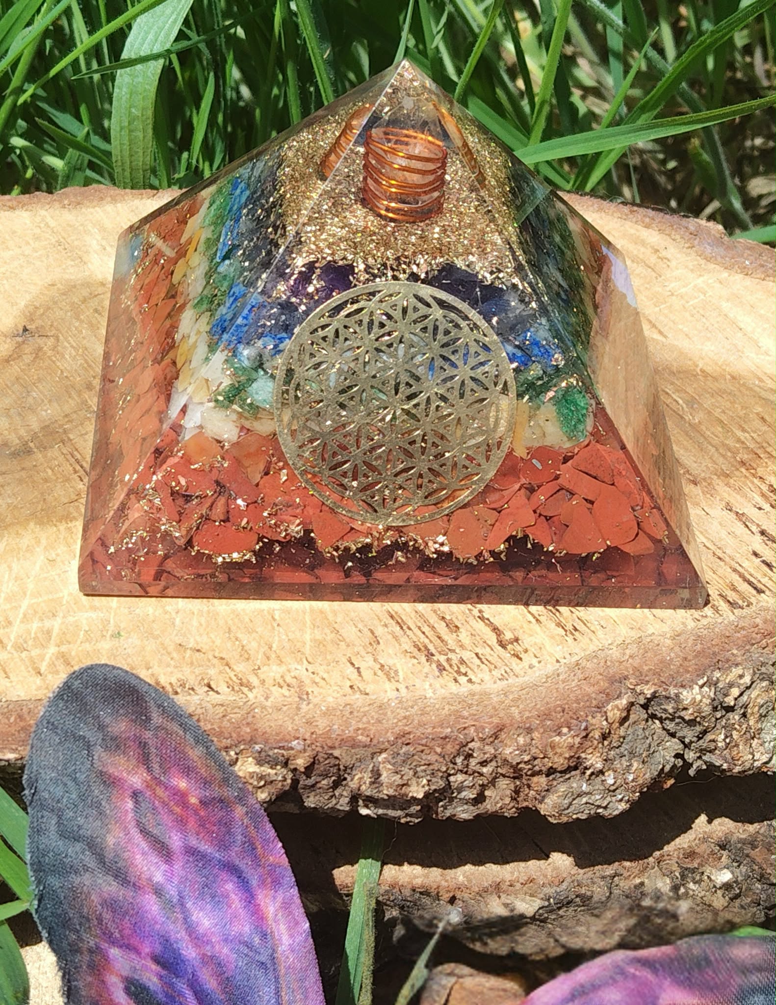 Pyramide orgonite 7 chakras avec fleur de vie et spirale de cuivre, posée sur une tranche de bois en extérieur (herbe, pierre violette au premier plan) ; évoque harmonie, protection, ancrage.
