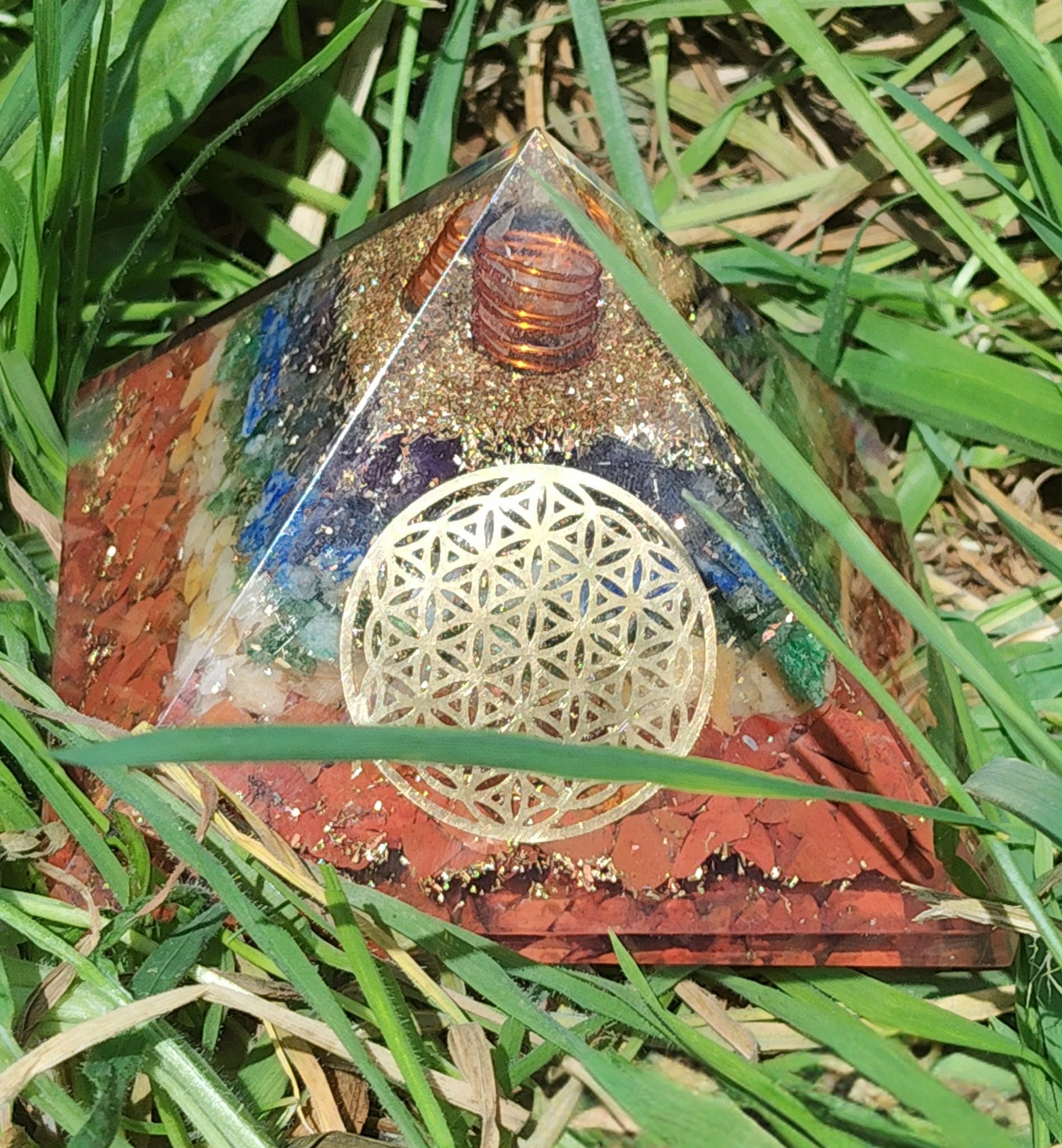 Pyramide orgonite 7 chakras avec fleur de vie et spirale de cuivre, posée dans l’herbe (photo nature) ; évoque harmonie, équilibre, protection.