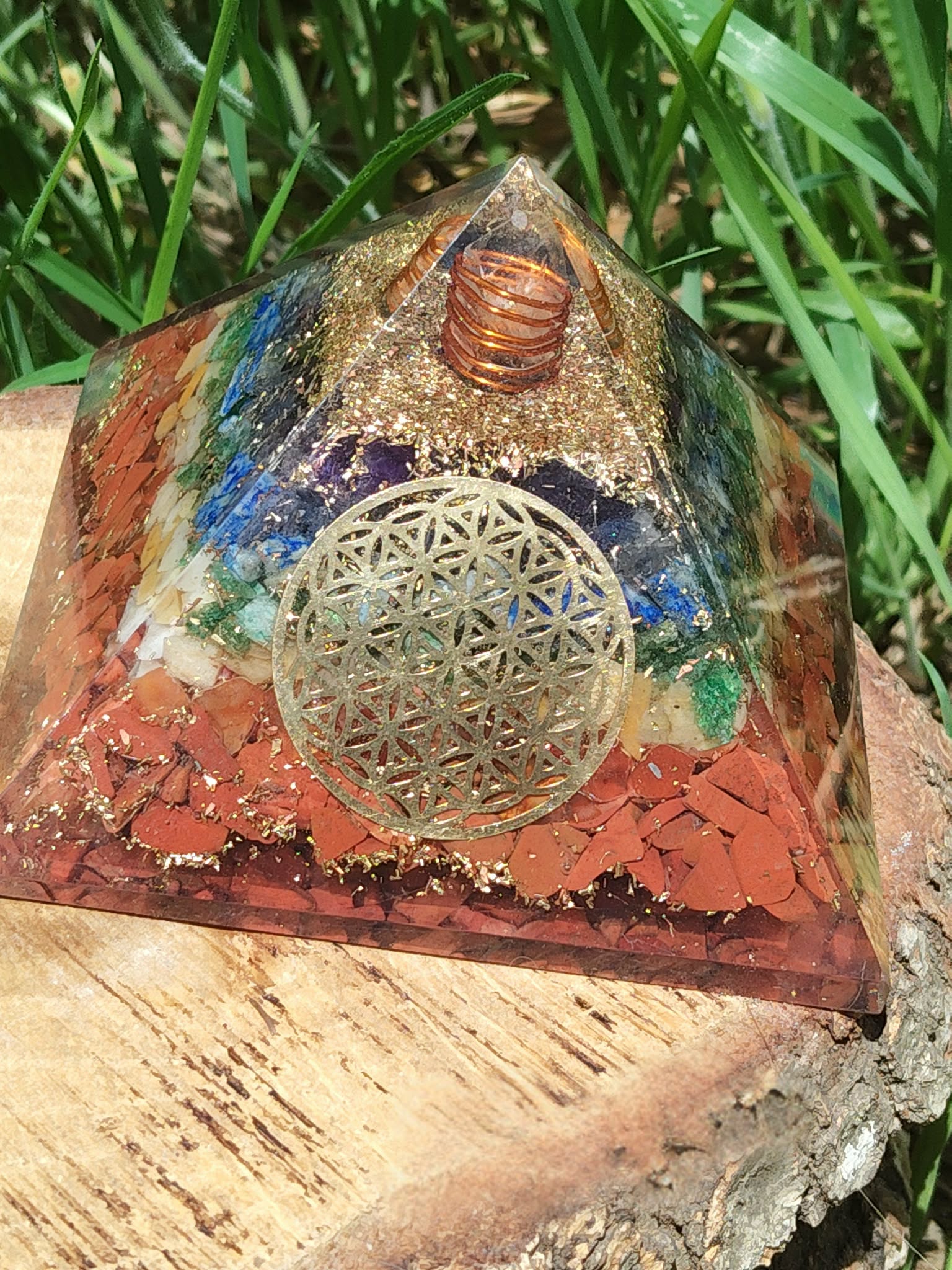 Pyramide orgonite 7 chakras avec fleur de vie et spirale de cuivre, posée sur tranche de bois en extérieur (herbe en arrière-plan) ; évoque équilibre, harmonisation, protection