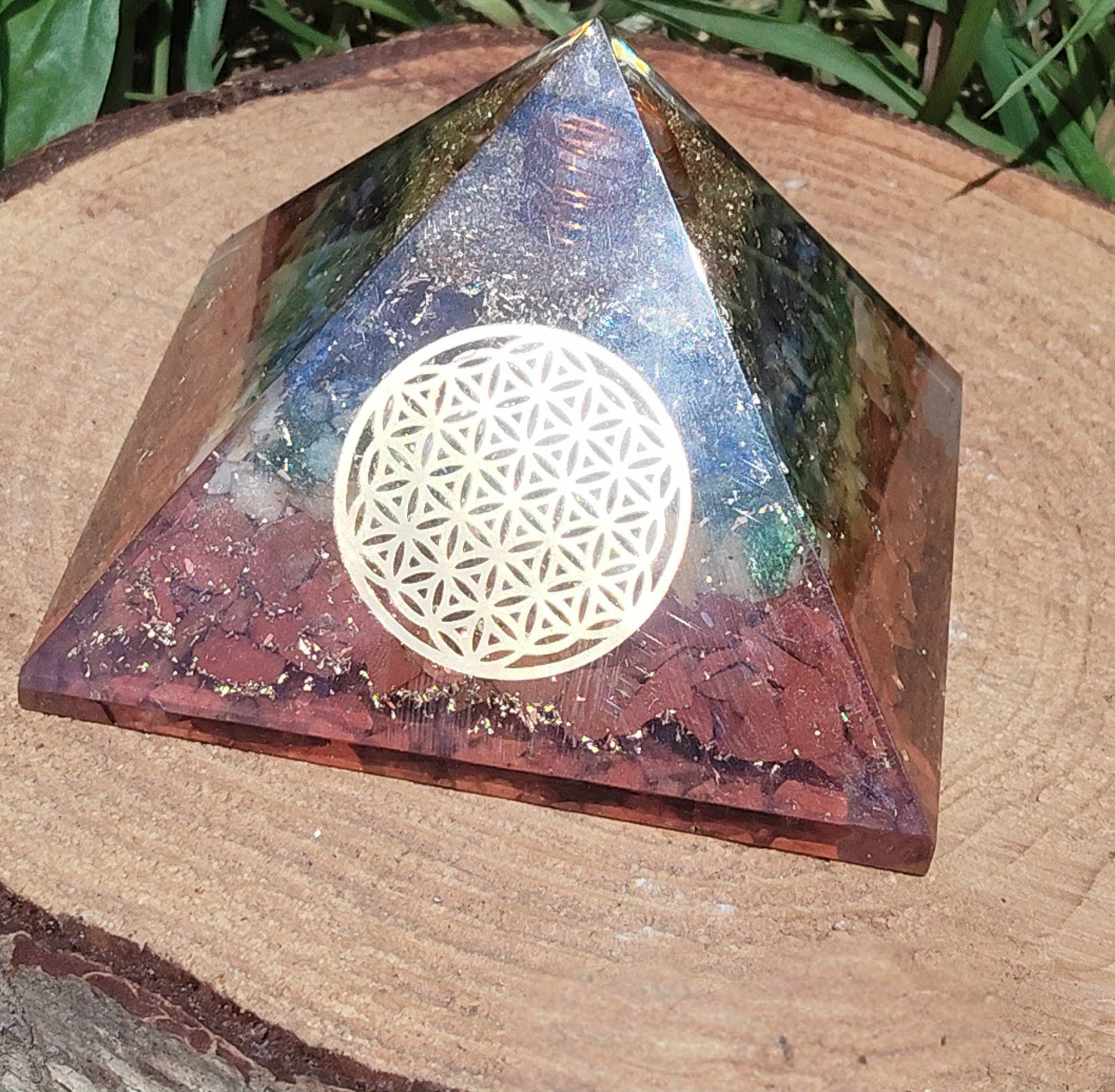 Pyramide orgonite 7 chakras avec fleur de vie (métal) et spirale de cuivre, posée sur tranche de bois en extérieur (fond végétal) ; évoque stabilité, recentrage, protection.