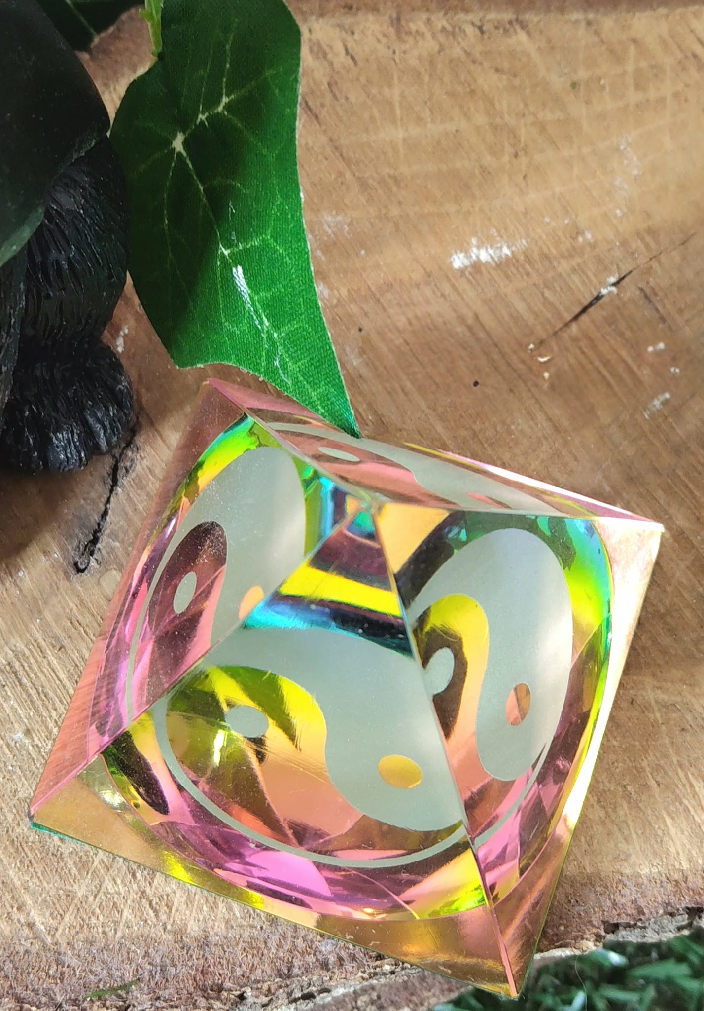 Pyramide en cristal irisé multicolore avec motif yin et yang, photographiée en biais sur une tranche de bois, avec décor vert et figurine sombre en arrière-plan ; inspire recentrage, équilibre émotionnel et atmosphère paisible.