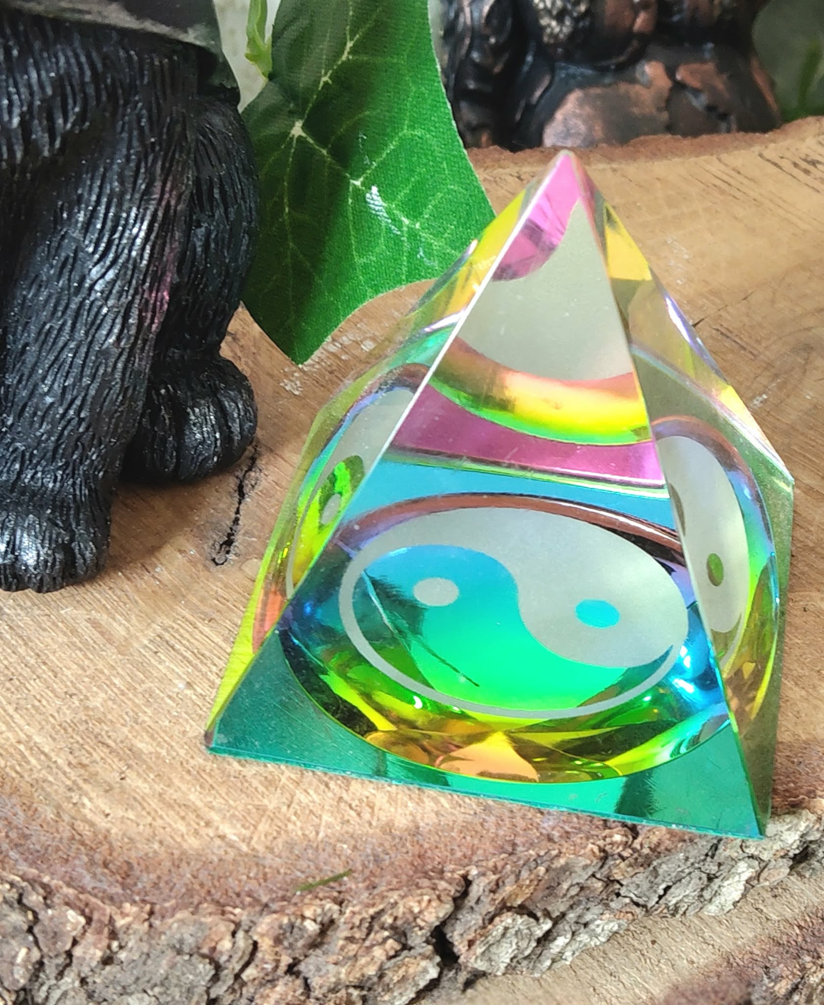 Pyramide en cristal irisé multicolore avec symbole yin et yang, posée sur une tranche de bois, avec une figurine noire et un décor vert flouté en arrière-plan ; aide à retrouver l’équilibre, calmer le mental et créer une ambiance harmonieuse.