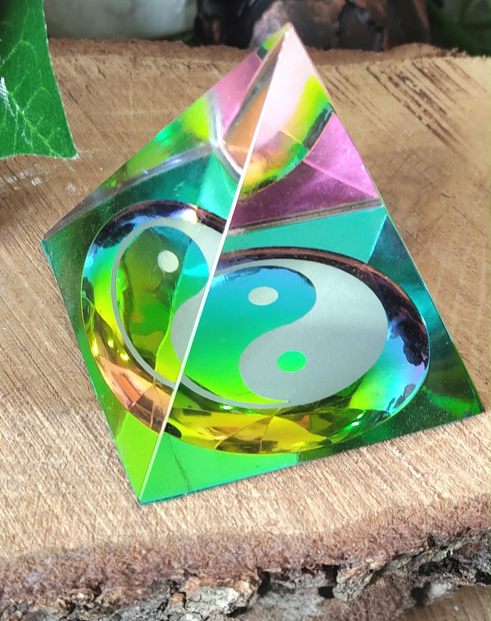 Pyramide en cristal irisé multicolore avec symbole yin et yang, posée sur une tranche de bois avec feuillage vert en arrière-plan ; invite à l’équilibre intérieur, la sérénité et l’harmonie des énergies.