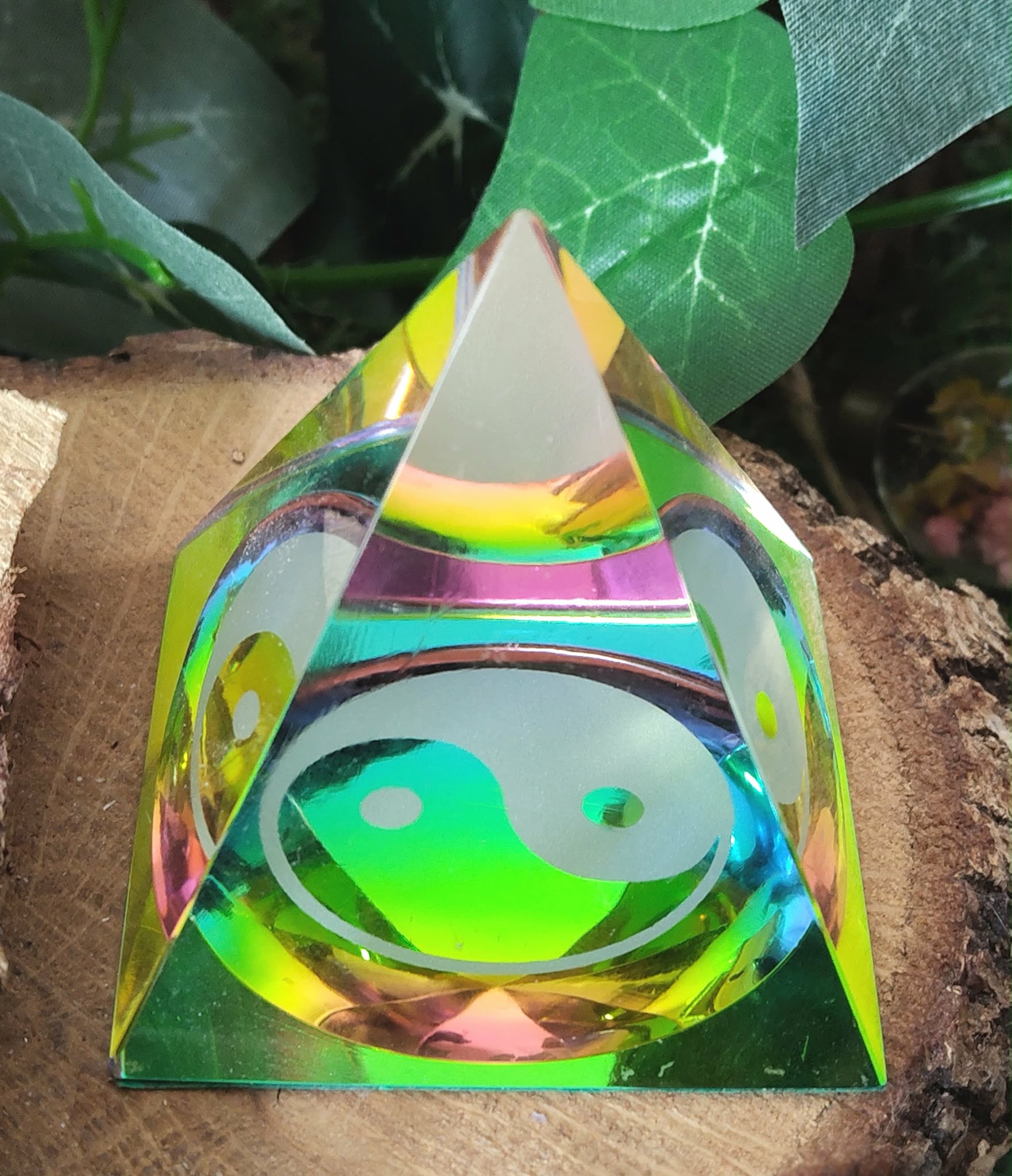 Pyramide en cristal irisé multicolore avec symbole yin et yang au centre, posée sur une tranche de bois, avec des feuilles vertes en arrière-plan ; apporte une ambiance d’équilibre, d’harmonie et d’apaisement.