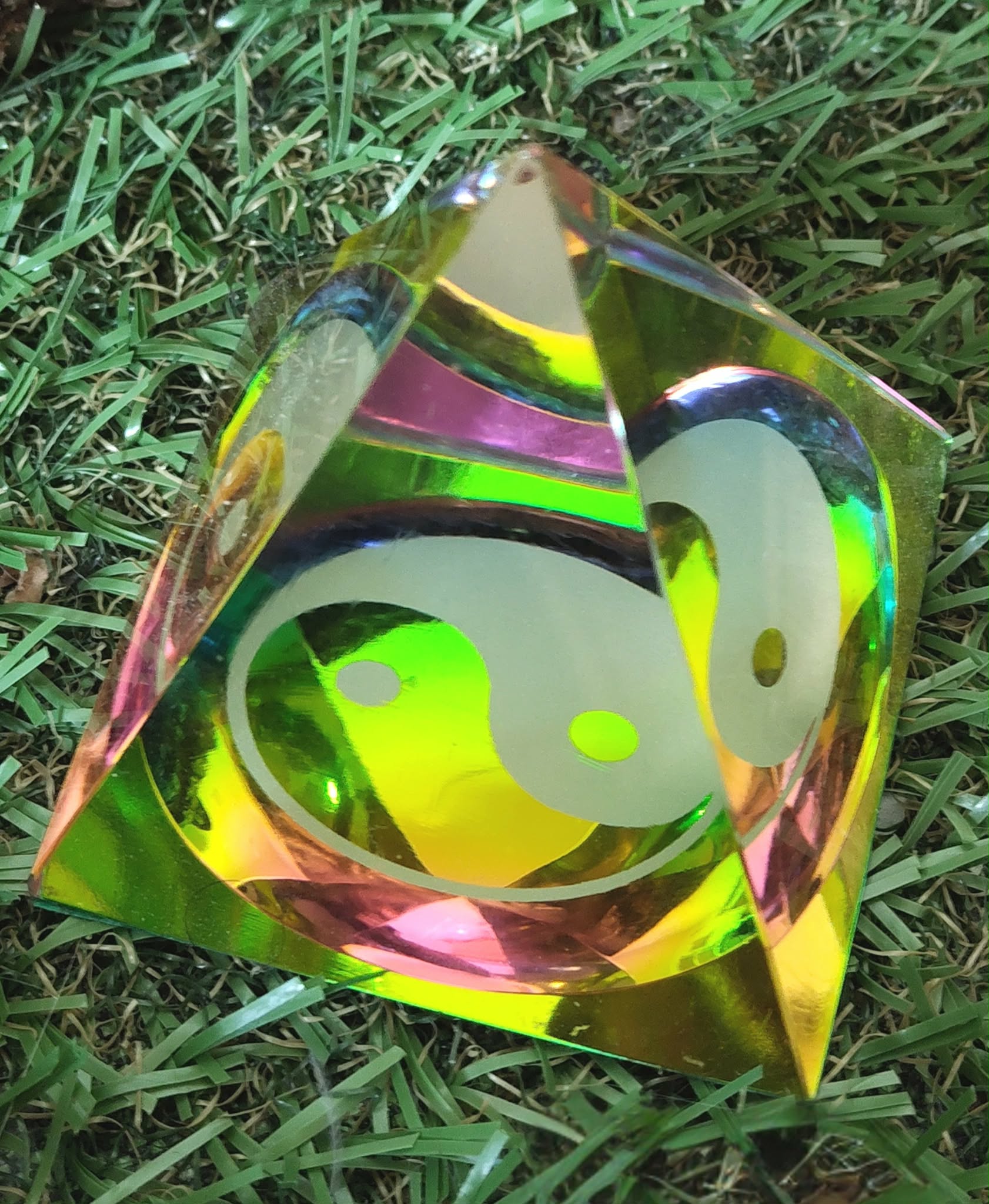 Pyramide en cristal irisé multicolore avec symbole yin et yang, posée sur un lit de feuillage vert ; apporte une sensation d’équilibre, d’harmonie et de calme intérieur.