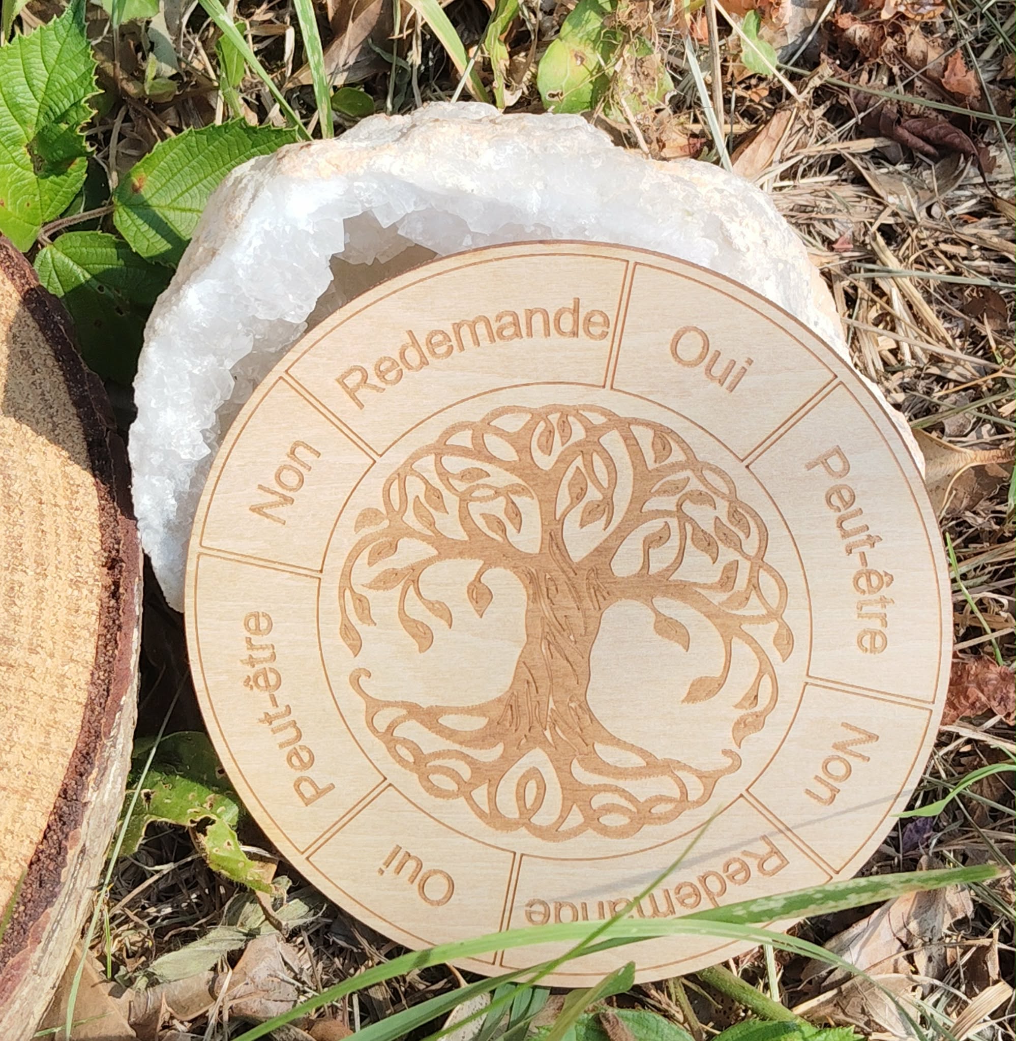 Plateau de pendule rond en bois gravé Arbre de Vie, posé dehors sur herbe et feuilles, repères Oui / Non / Peut-être / Redemande autour.