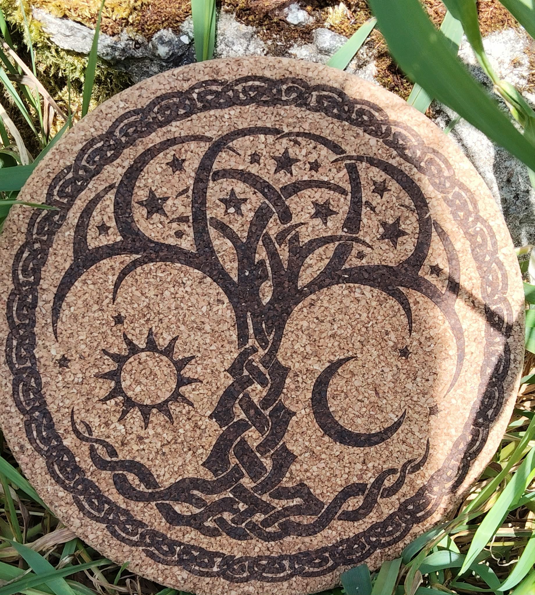 Dessous de plat rond en liège gravé arbre de vie avec soleil, lune et étoiles, bordure celtique; photo en extérieur dans l’herbe.