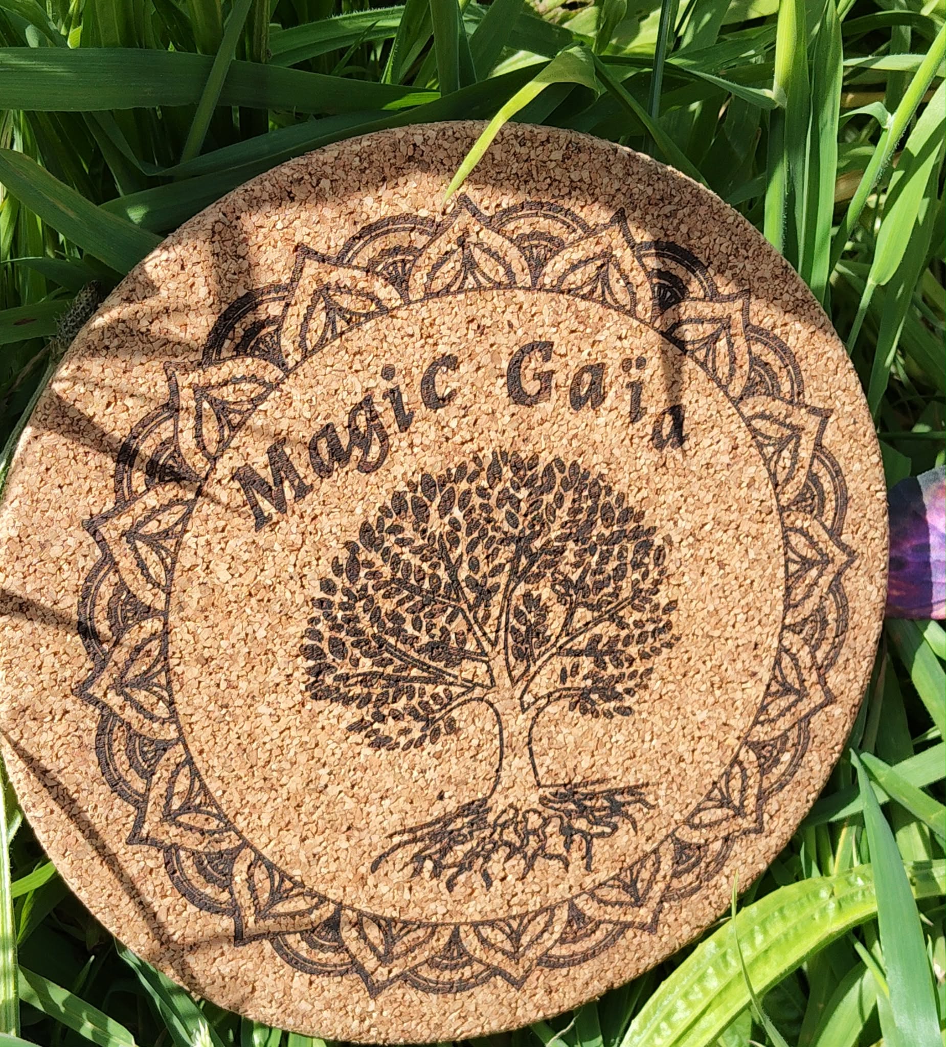 Dessous de plat rond en liège gravé “Magic Gaïa” avec arbre central et bordure mandala; photo en extérieur dans l’herbe, ombres de feuilles.