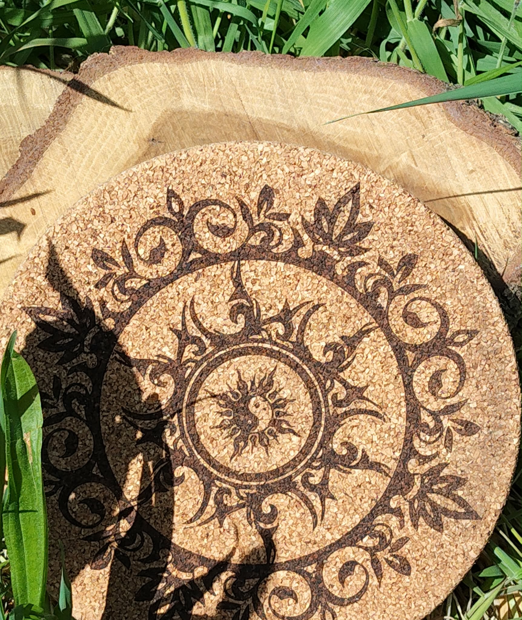 Dessous de plat rond en liège gravé d’un soleil et d’un croissant de lune, entourés de motifs végétaux; photo en extérieur dans l’herbe.
