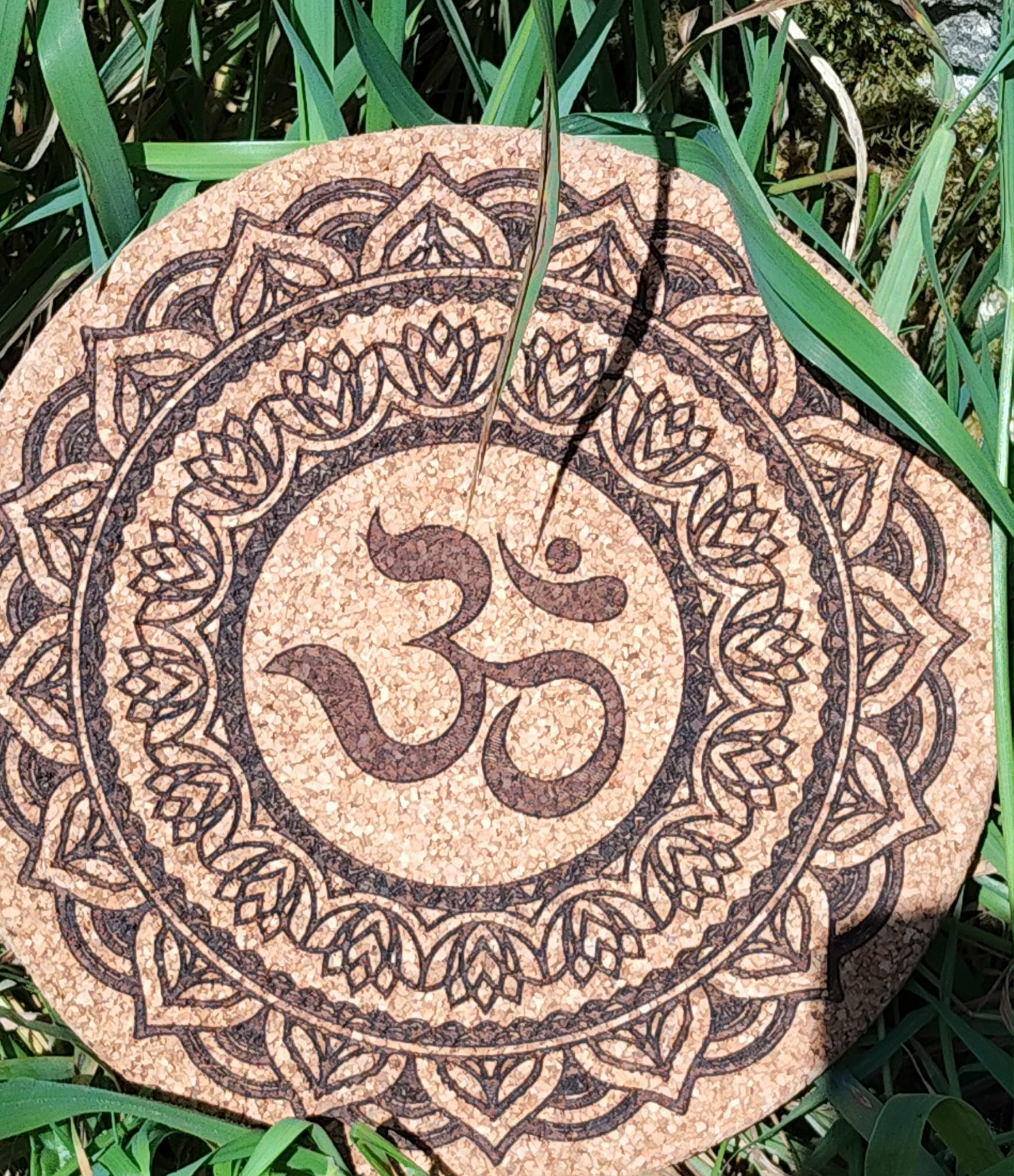 Dessous de plat rond en liège gravé symbole Om au centre, entouré d’un mandala et motifs géométriques; photo en extérieur dans l’herbe.