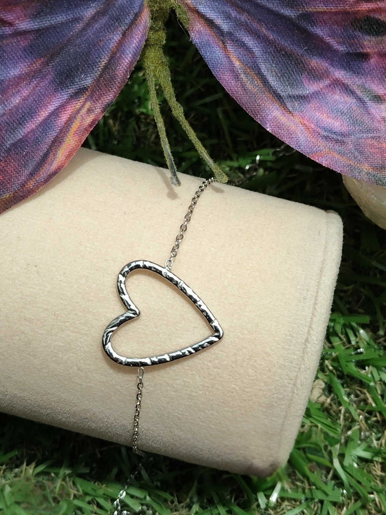Bracelet chaîne fine en acier inoxydable, pendentif cœur ajouré; posé sur support blanc en extérieur, herbe en fond et papillon violet.