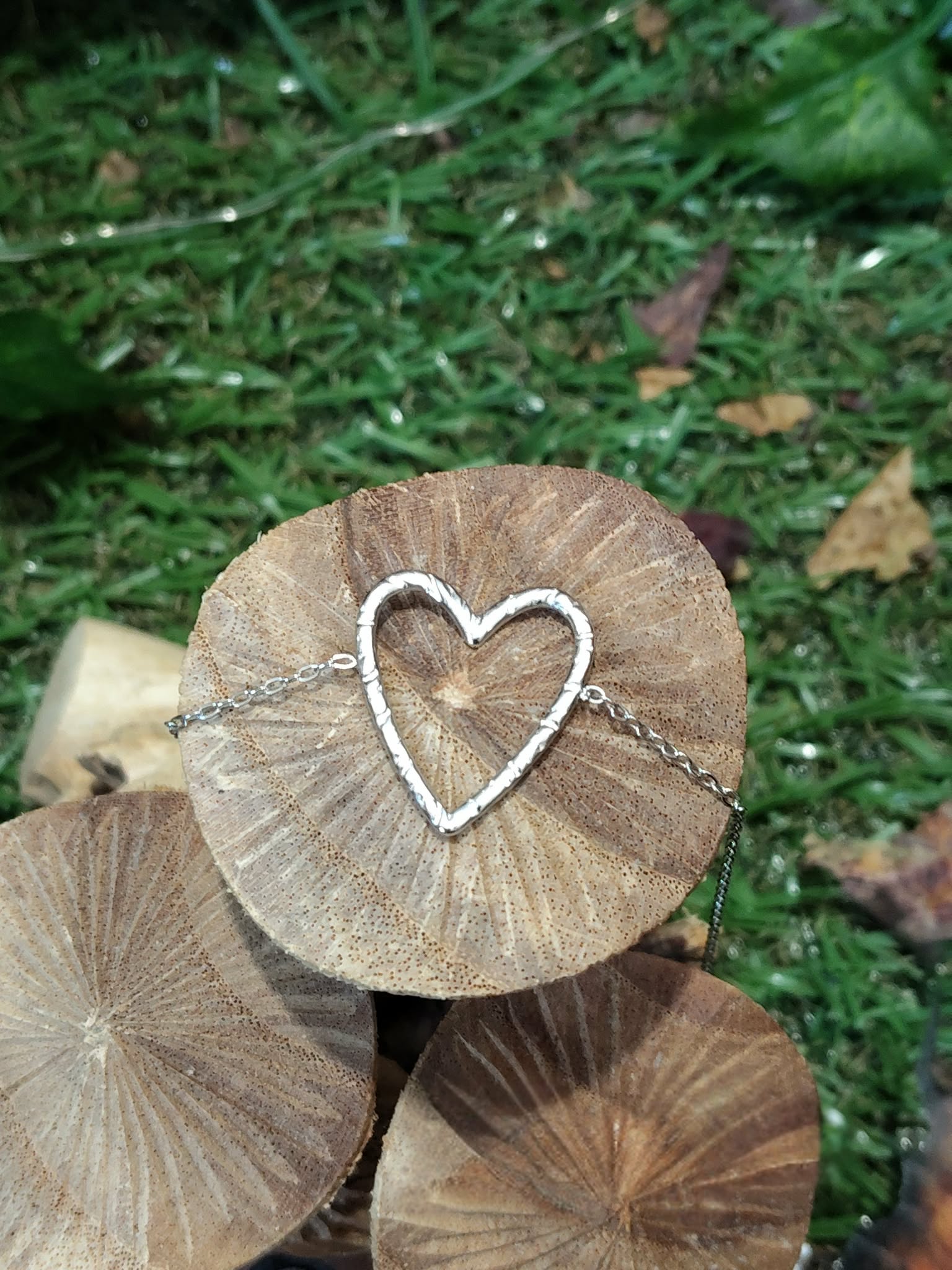 Bracelet chaîne fine en acier inoxydable, pendentif cœur ajouré; posé sur rondins de bois, décor mousse et feuilles en extérieur.