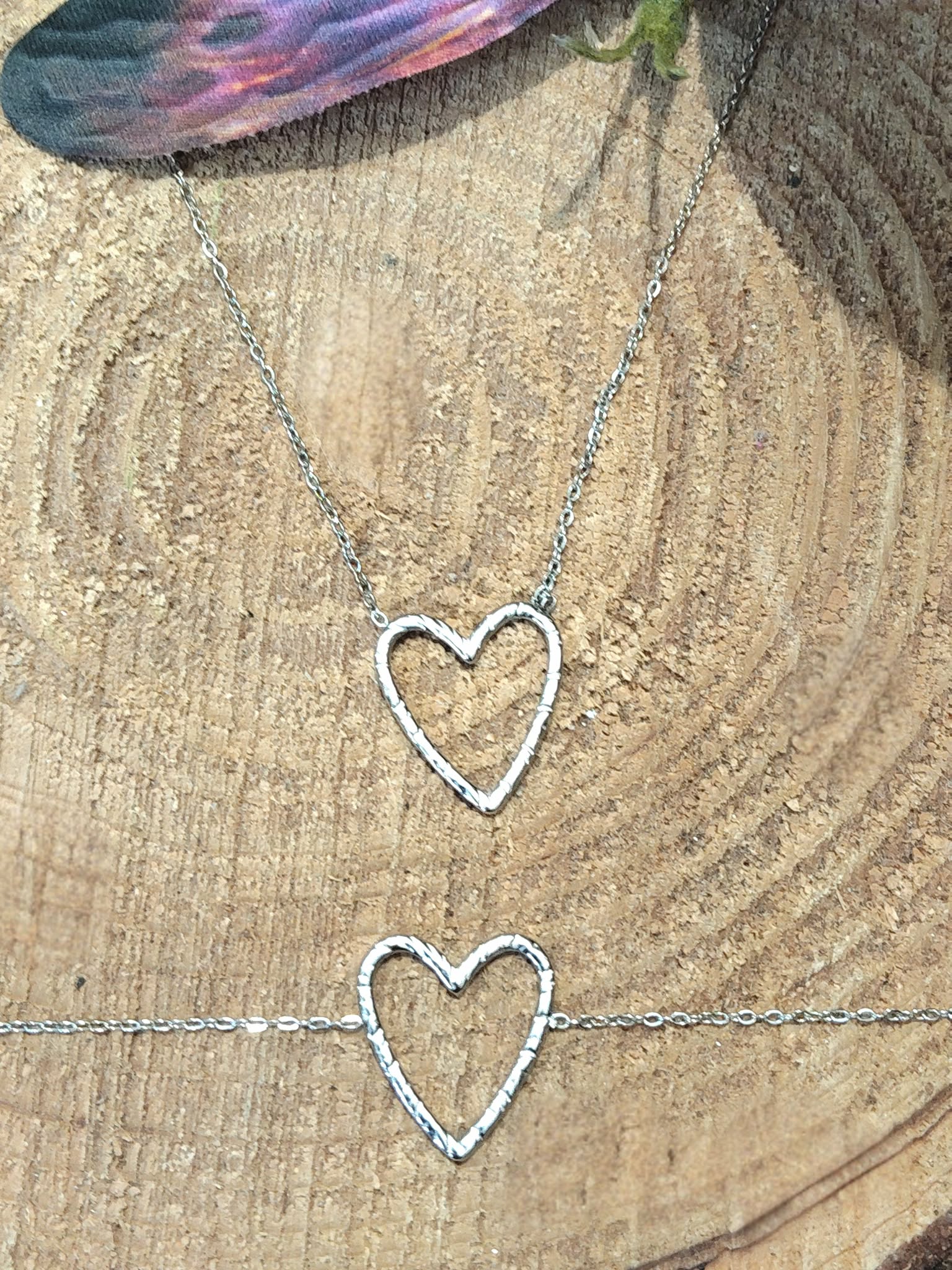 Deux bracelets chaîne fine en acier inoxydable avec pendentif cœur ajouré; posés sur rondin de bois, décor nature en extérieur.