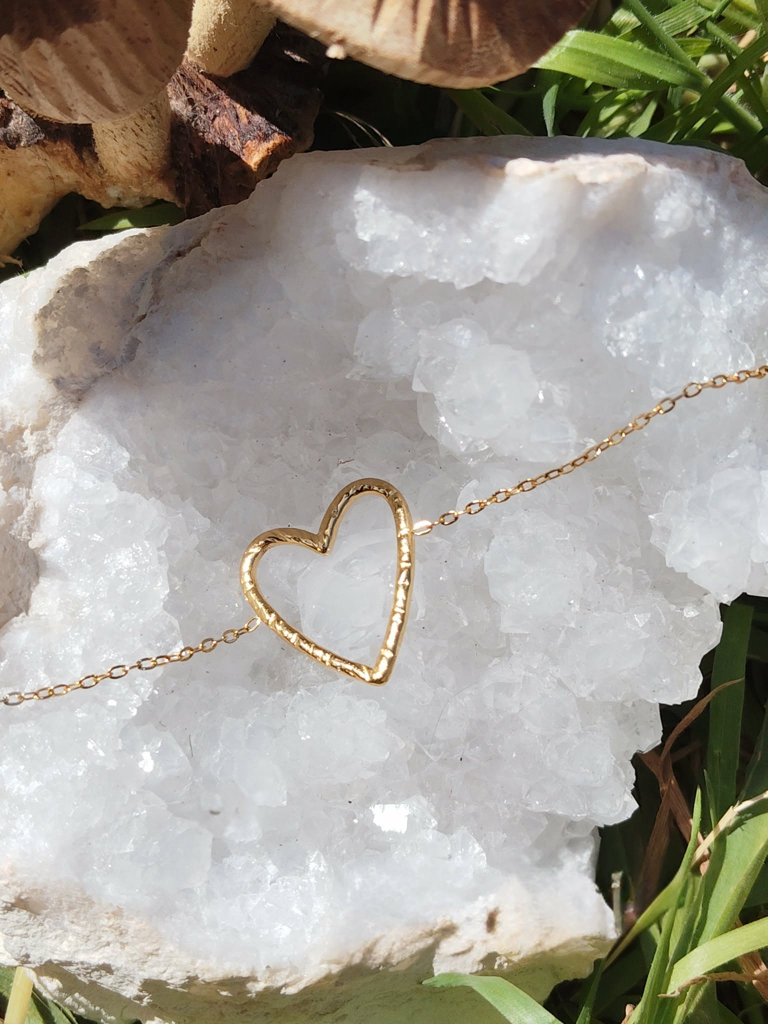 Bracelet chaîne fine en acier inoxydable doré, pendentif cœur ajouré; posé sur pierre blanche en extérieur, décor nature en arrière-plan.