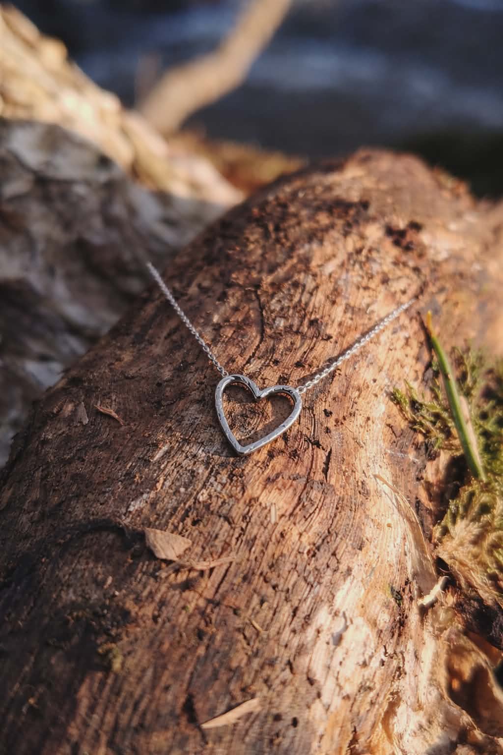 Pendentif cœur ajouré martelé en acier inoxydable argenté sur chaîne fine, posé sur tronc d’arbre avec écorce et mousse, bijou lumineux au style naturel
