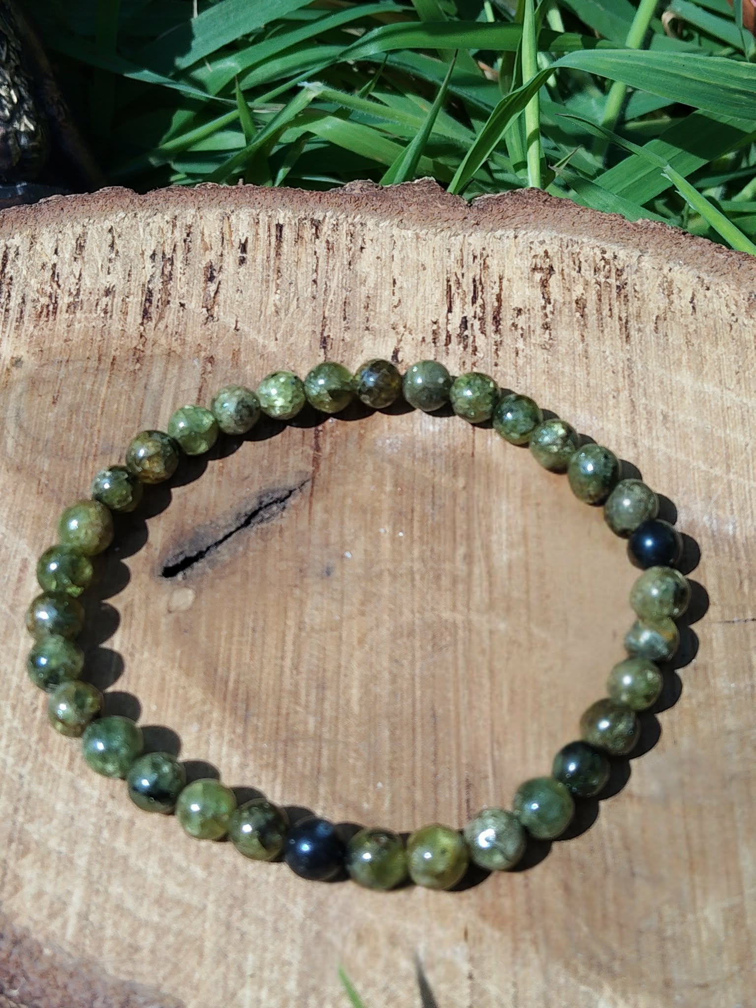 Bracelet péridot vert olive sur tranche de bois avec herbe en fond, perles naturelles pour apaisement du mental, renouveau et stabilité du ressenti