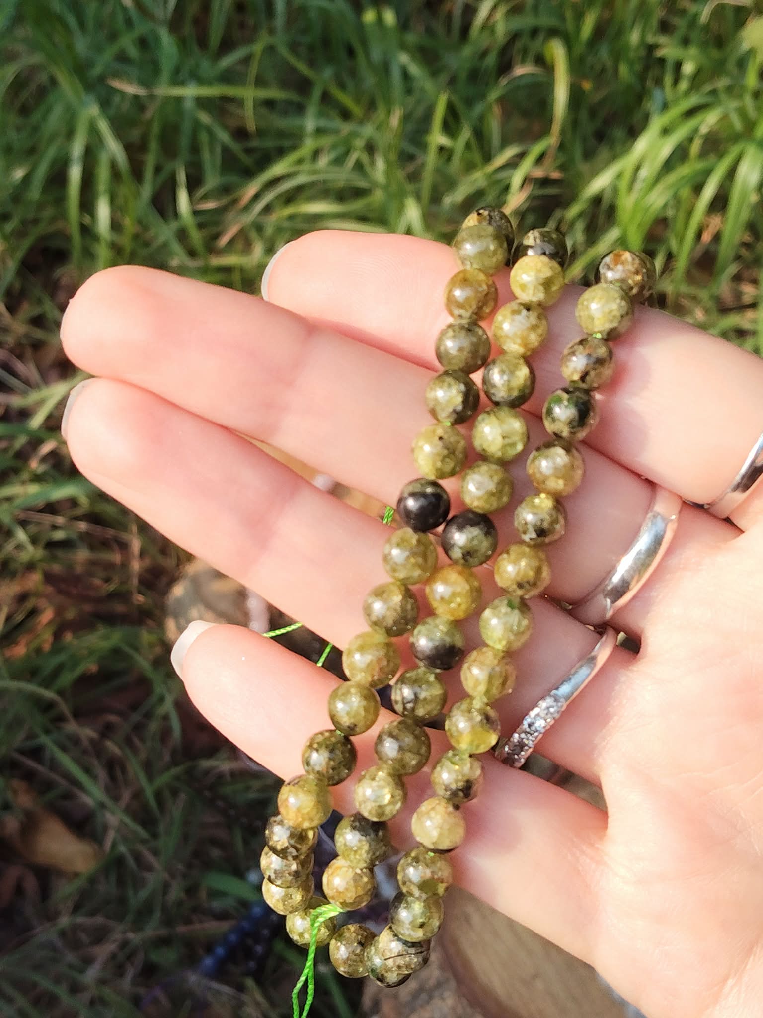 Fil de perles de péridot tenu en main sur fond d’herbe verte, nuances vert marbré et noir pour énergie lumineuse, renouveau et élan intérieur