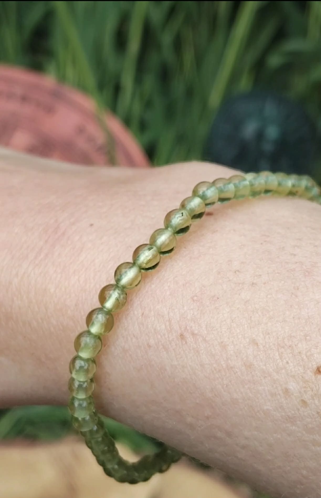 Bracelet en péridot (perles 4 mm vert olive translucide), monté sur fil, porté au poignet ; photo en extérieur avec feuillage vert en arrière-plan, ambiance fraîche et lumineuse, évoque vitalité, optimisme et légèreté.