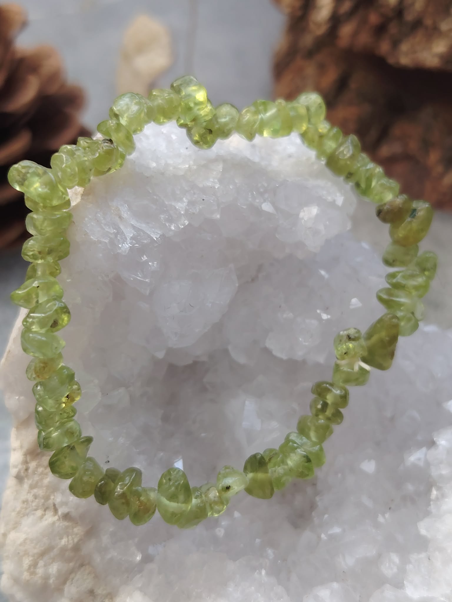 Bracelet péridot baroque (chips vert olive translucides) formant un cercle autour d’une géode blanche, posé sur un décor naturel (mousse/bois) avec arrière-plan doux, photo en gros plan.