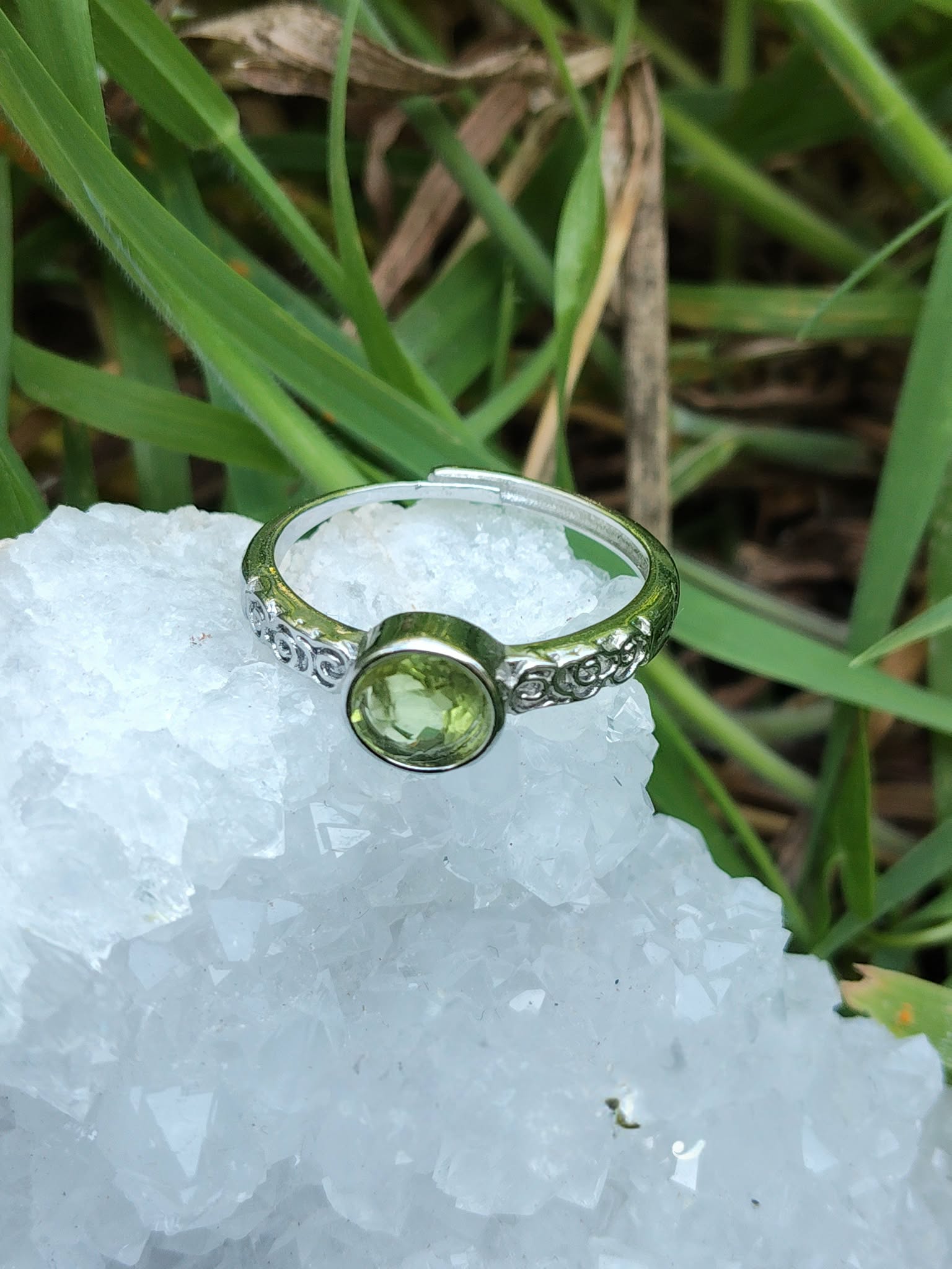 Bague péridot facetté réglable en cuivre blanc, posée sur une géode blanche, herbes vertes en arrière-plan ; apporte apaisement et clarté