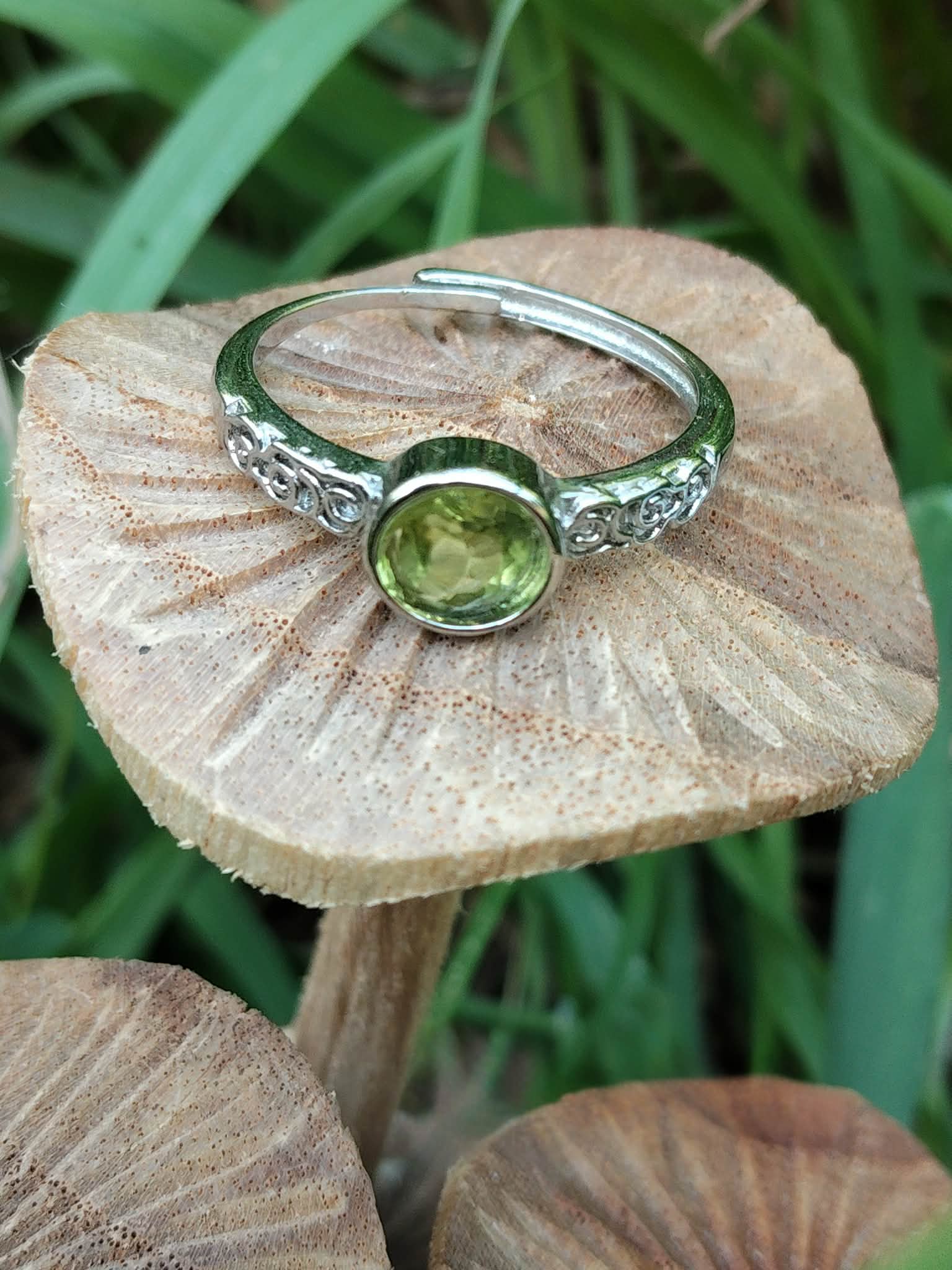 Bague réglable en cuivre blanc, péridot facetté vert rond, posée sur champignon en bois, fond de feuilles vertes ; évoque la joie, la clarté mentale, l’élan positif et l’équilibre émotionnel.