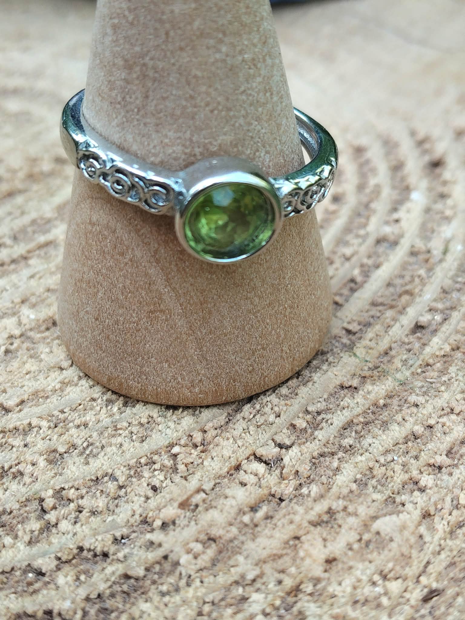Bague réglable en cuivre blanc avec péridot facetté vert, posée sur un petit support en bois sur une rondelle de tronc ; apporte clarté, optimisme et apaisement.