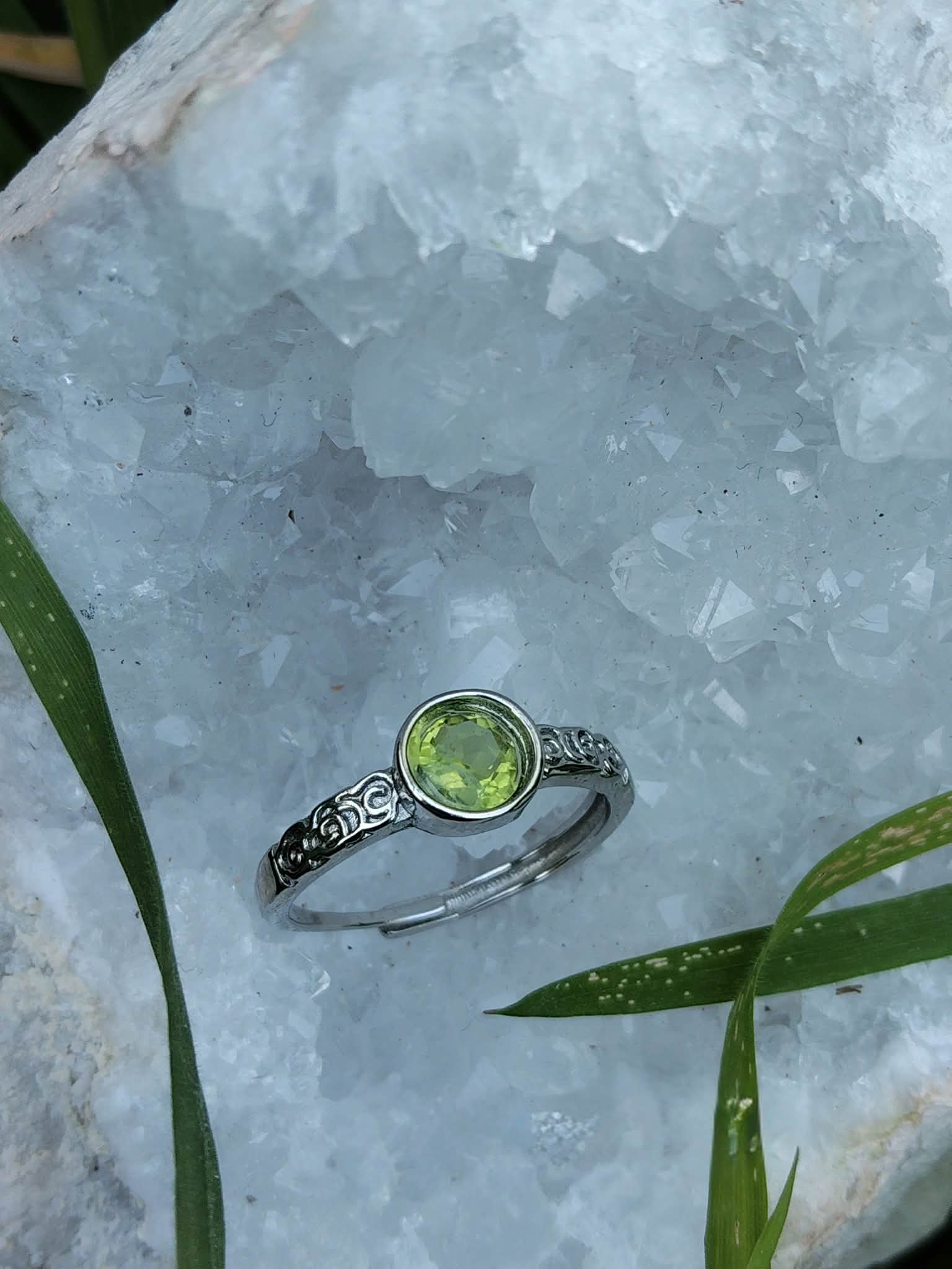 Bague réglable en cuivre blanc avec péridot facetté, posée sur une géode blanche cristallisée ; aide à retrouver optimisme, clarté mentale et élan positif