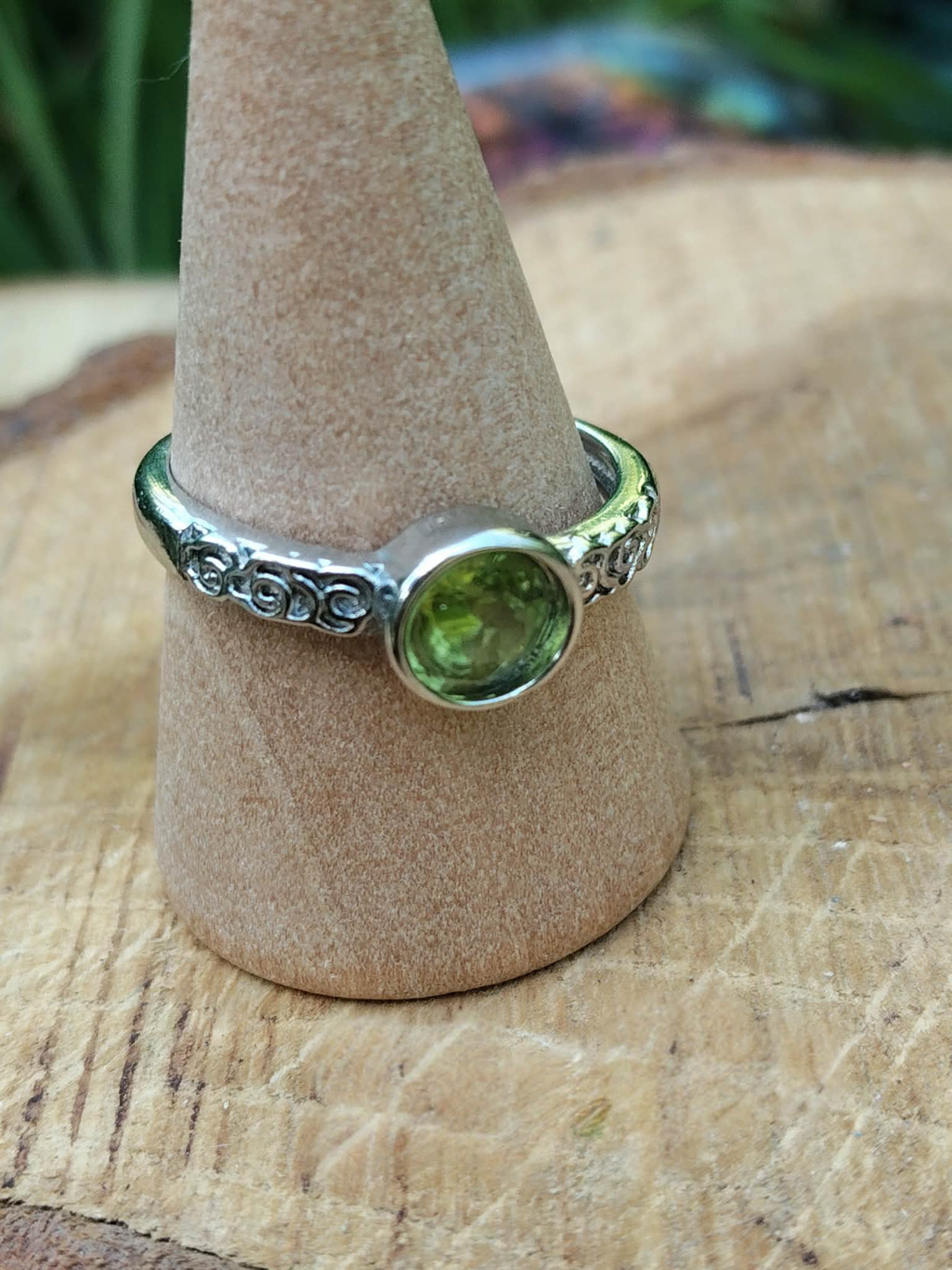 Bague réglable en cuivre blanc avec péridot facetté rond vert, portée sur un doigt, prise de vue rapprochée sur fond bois clair ; associée à l’optimisme, à la clarté d’esprit et à l’apaisement émotionnel.