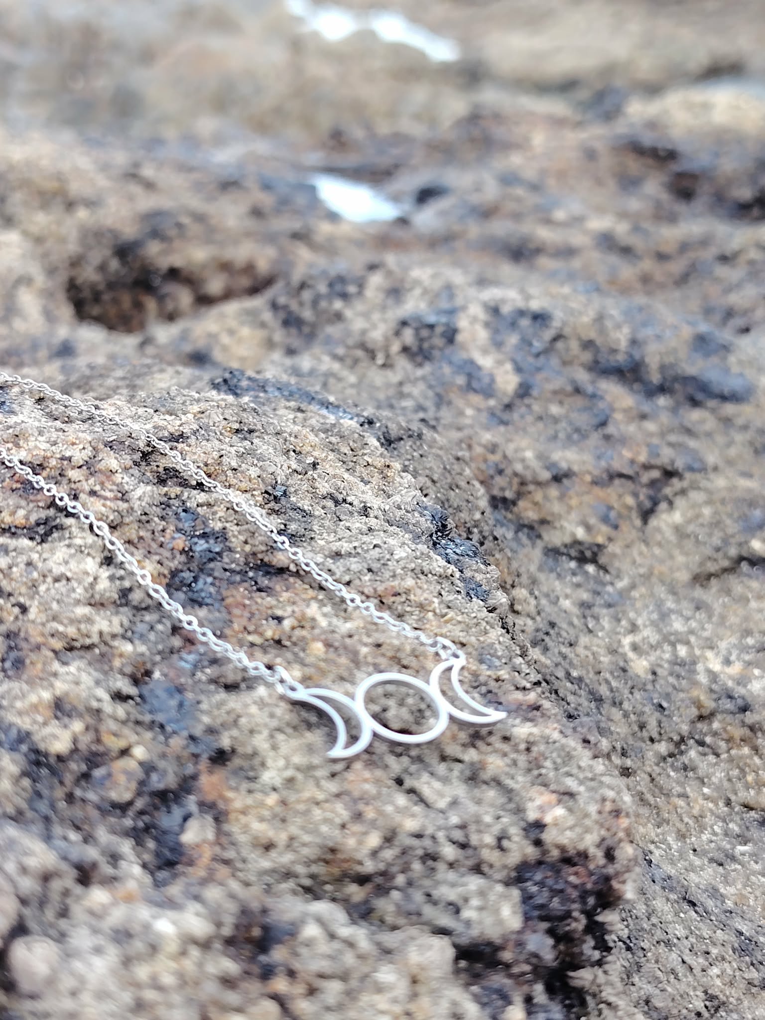 Collier fin triple lune argenté posé sur rocher granit en décor minéral sauvage, symbole des phases lunaires en Bretagne