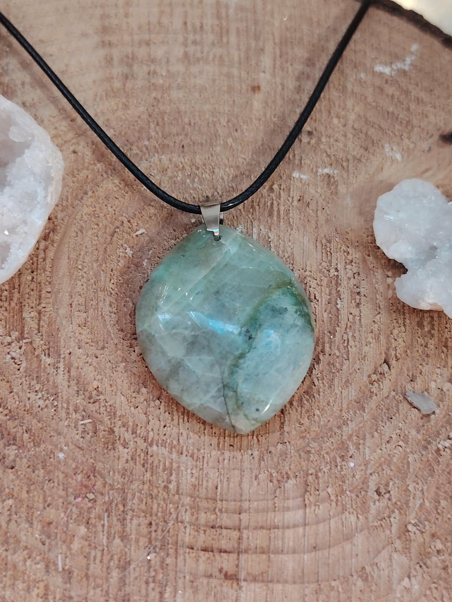 Pendentif en garniérite vert-bleuté sur cordon noir, pierre polie posée sur bois avec petits galets blancs, bijou en pierre naturelle évoquant le recul, la sérénité et l’équilibre intérieur.
