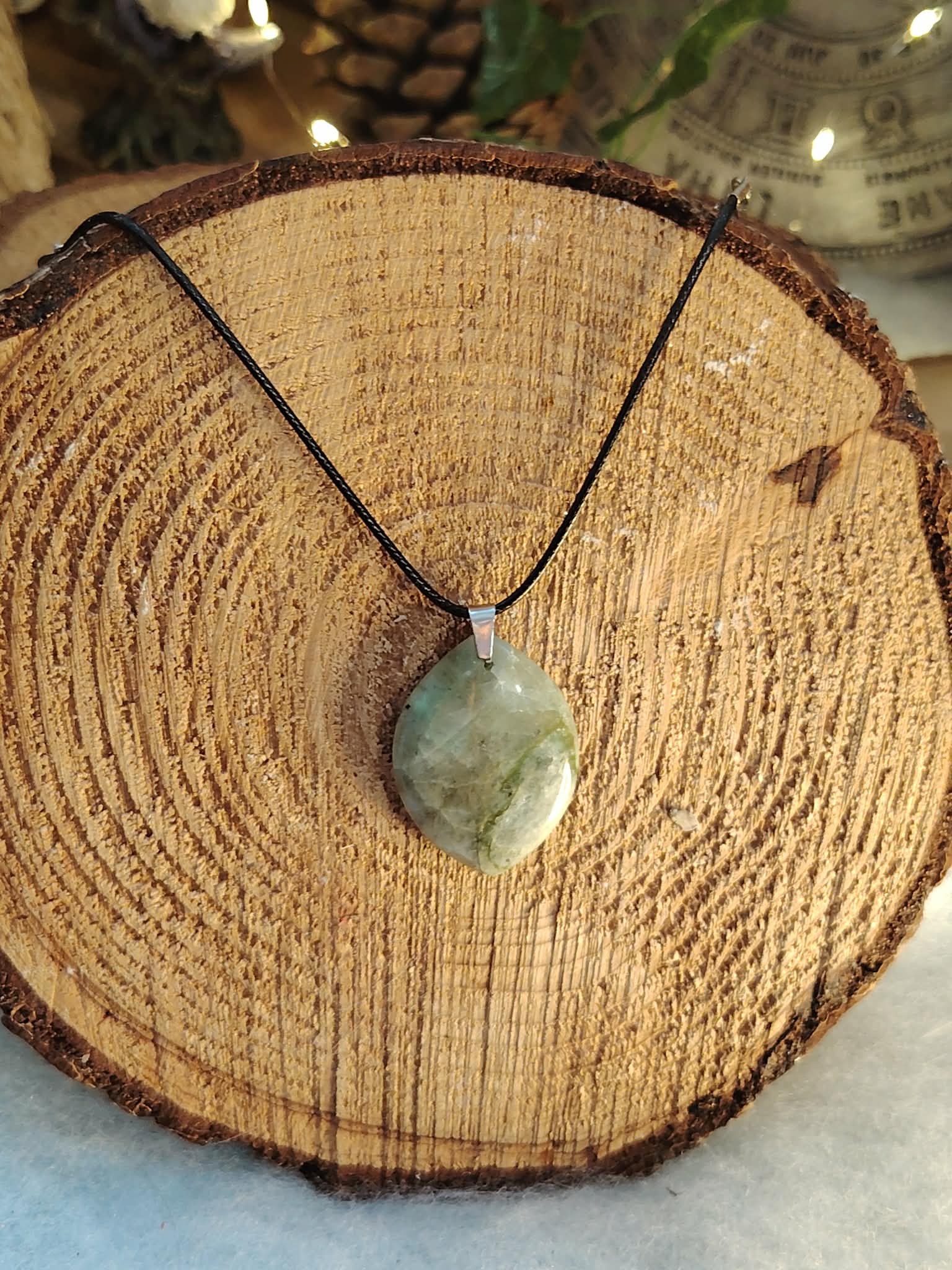 Pendentif garniérite vert-gris facetté sur cordon noir avec bélière argentée, posé au centre d’une tranche de bois aux cernes visibles, pierre naturelle associée à la douceur, à la stabilité et à l’harmonie émotionnelle.