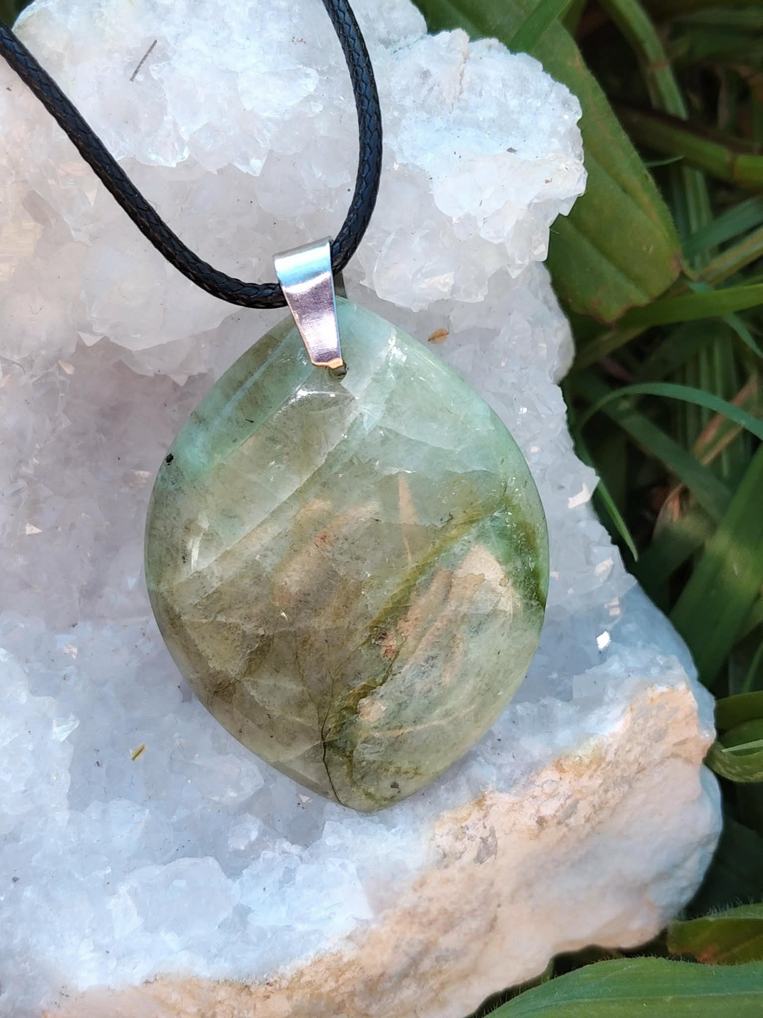 Pendentif en garniérite vert pâle sur cordon noir avec bélière argentée, posé sur géode blanche et herbes vertes, pierre naturelle associée à la douceur, au recentrage et à l’équilibre intérieur.