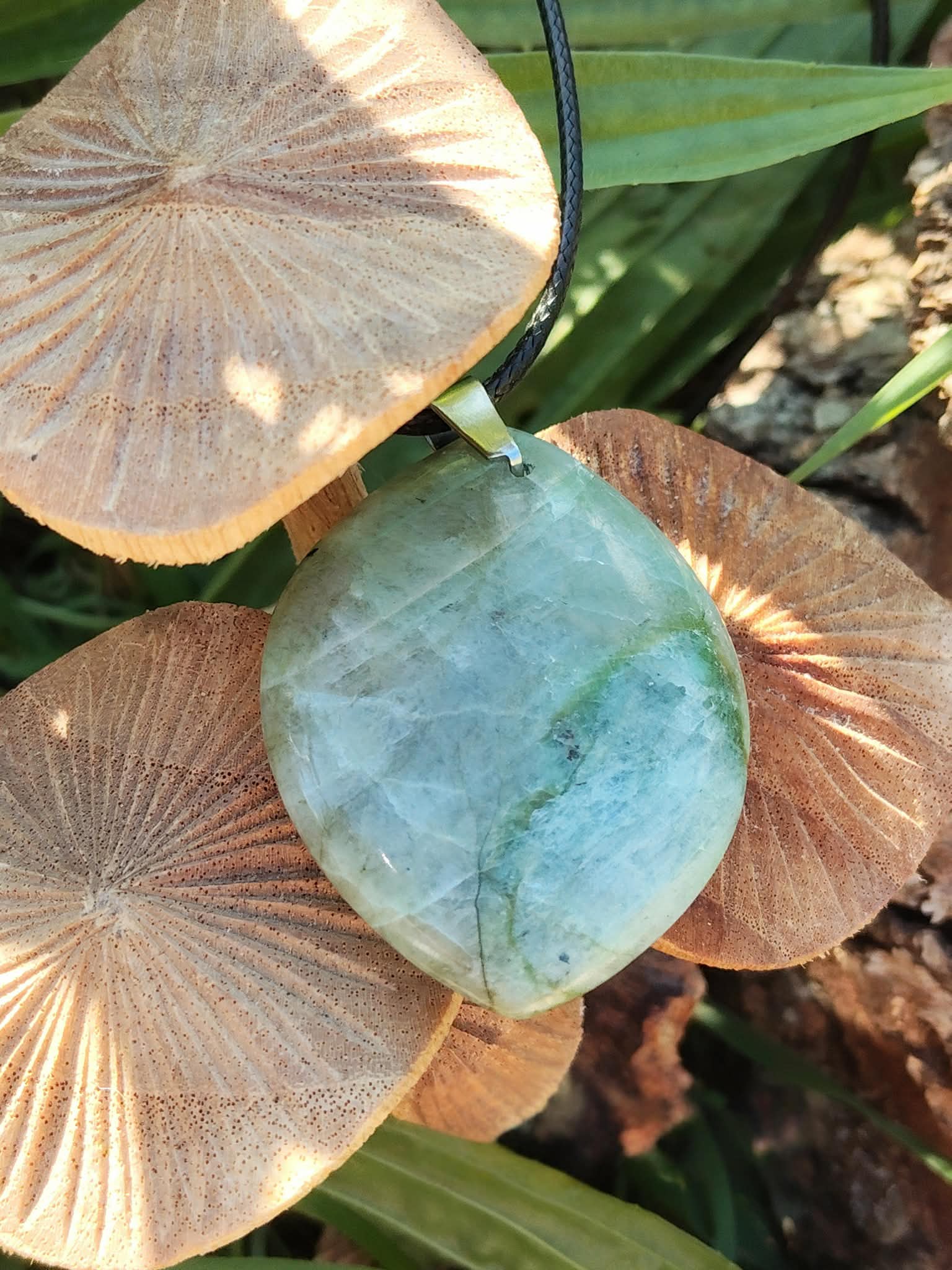 Pendentif en garniérite vert d’eau sur cordon noir, posé sur champignons en bois et feuilles vertes en décor sous-bois, pierre naturelle évoquant l’harmonie du cœur, la paix intérieure et l’ancrage doux.