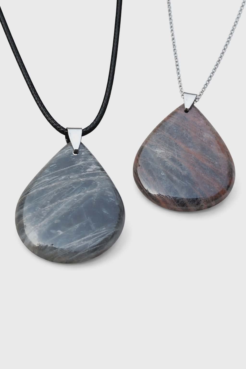 Deux pendentifs goutte en pierre de lune noire polie, l’un sur cordon noir et l’autre sur chaîne argentée, photo détourée sur fond blanc, bijoux en pierre naturelle reliés à l’apaisement, à la stabilité et à la sérénité intérieure.
