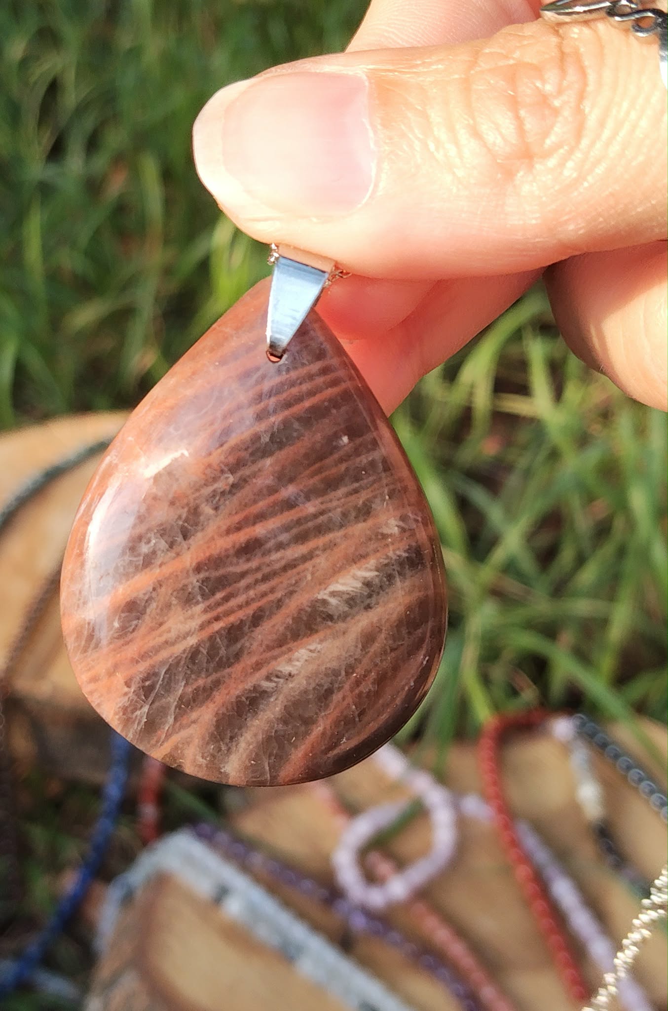 Pendentif goutte en pierre de lune noire polie tenu entre les doigts, avec herbe et feuillage en arrière-plan, bijou en pierre naturelle évoquant la sensibilité maîtrisée, l’équilibre du cœur et la présence à soi.