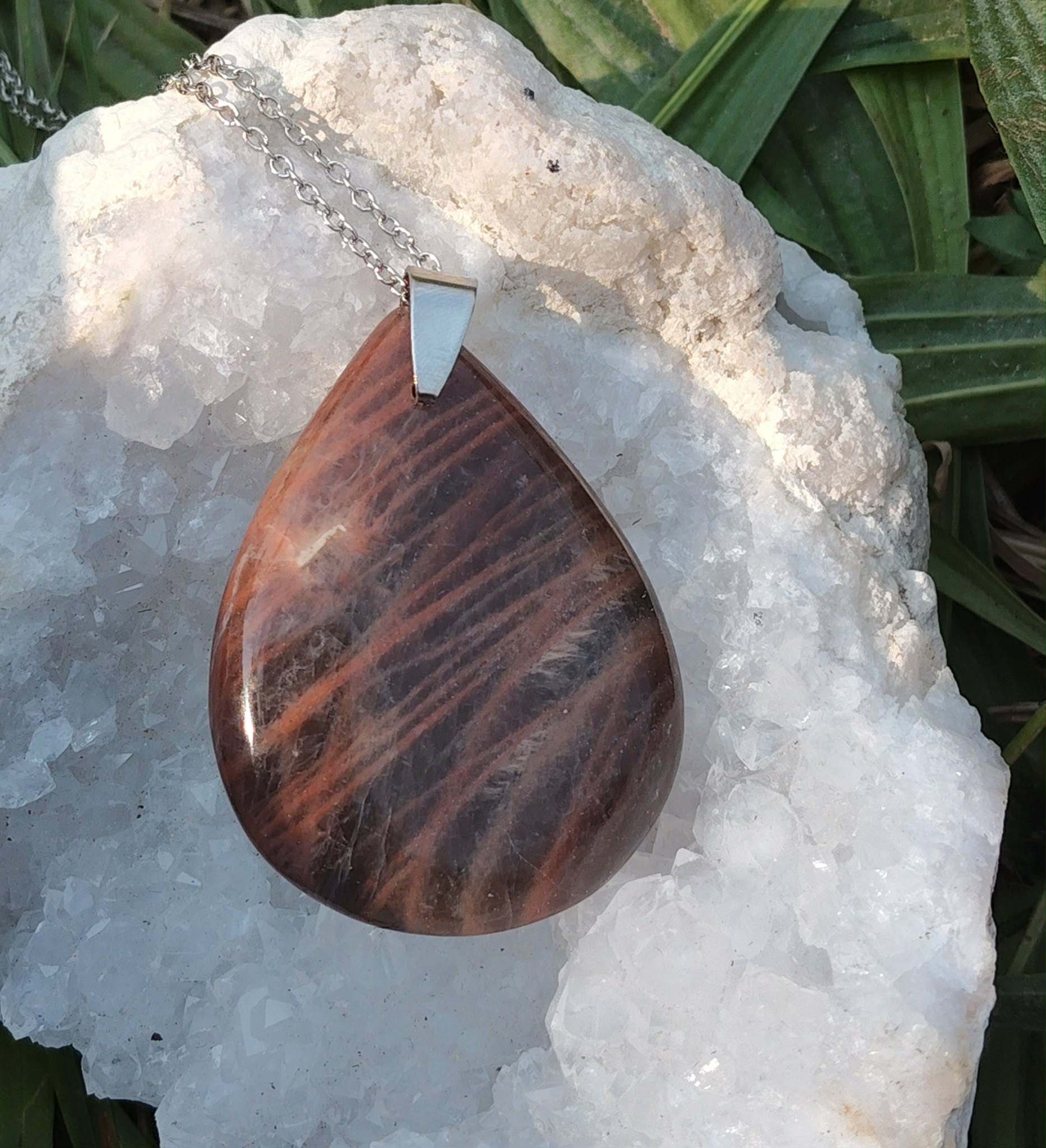 Pendentif goutte en pierre de lune noire polie sur chaîne argentée, posé sur une géode blanche avec feuillage en arrière-plan, bijou en pierre naturelle lié à l’apaisement, à l’intuition calme et à la stabilité intérieure.