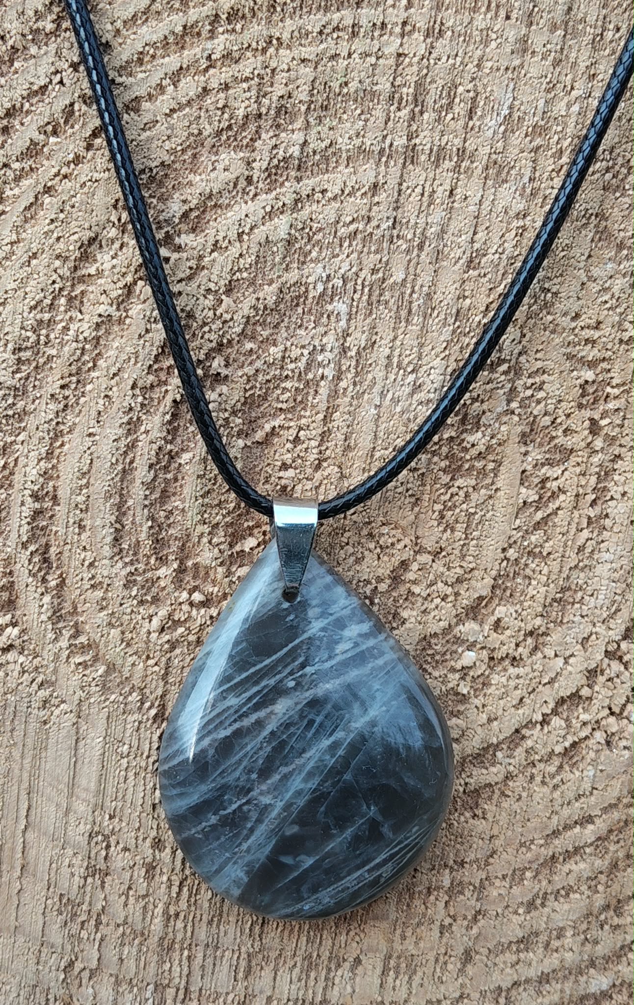 Pendentif goutte en pierre de lune noire polie sur cordon noir, posé sur une tranche de bois, pierre naturelle associée au recentrage, à la clarté émotionnelle et à la tranquillité intérieure.