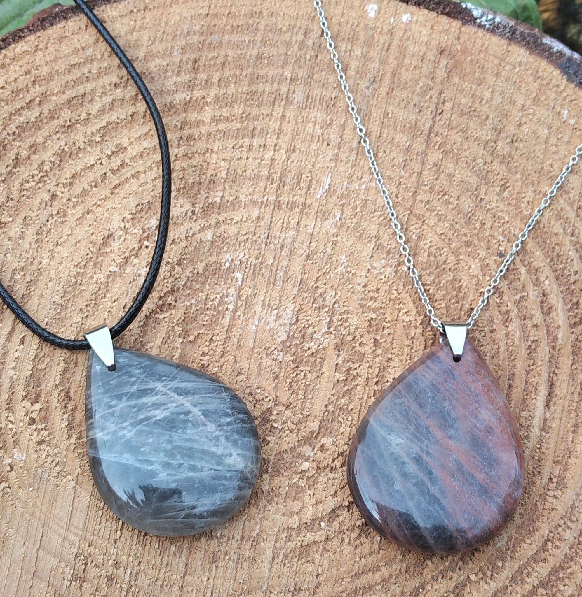 Deux pendentifs goutte en pierre de lune noire polie, l’un sur cordon noir et l’autre sur chaîne argentée, posés sur une tranche de bois, bijoux en pierre naturelle associés à la paix intérieure, à l’ancrage et à l’harmonie émotionnelle.