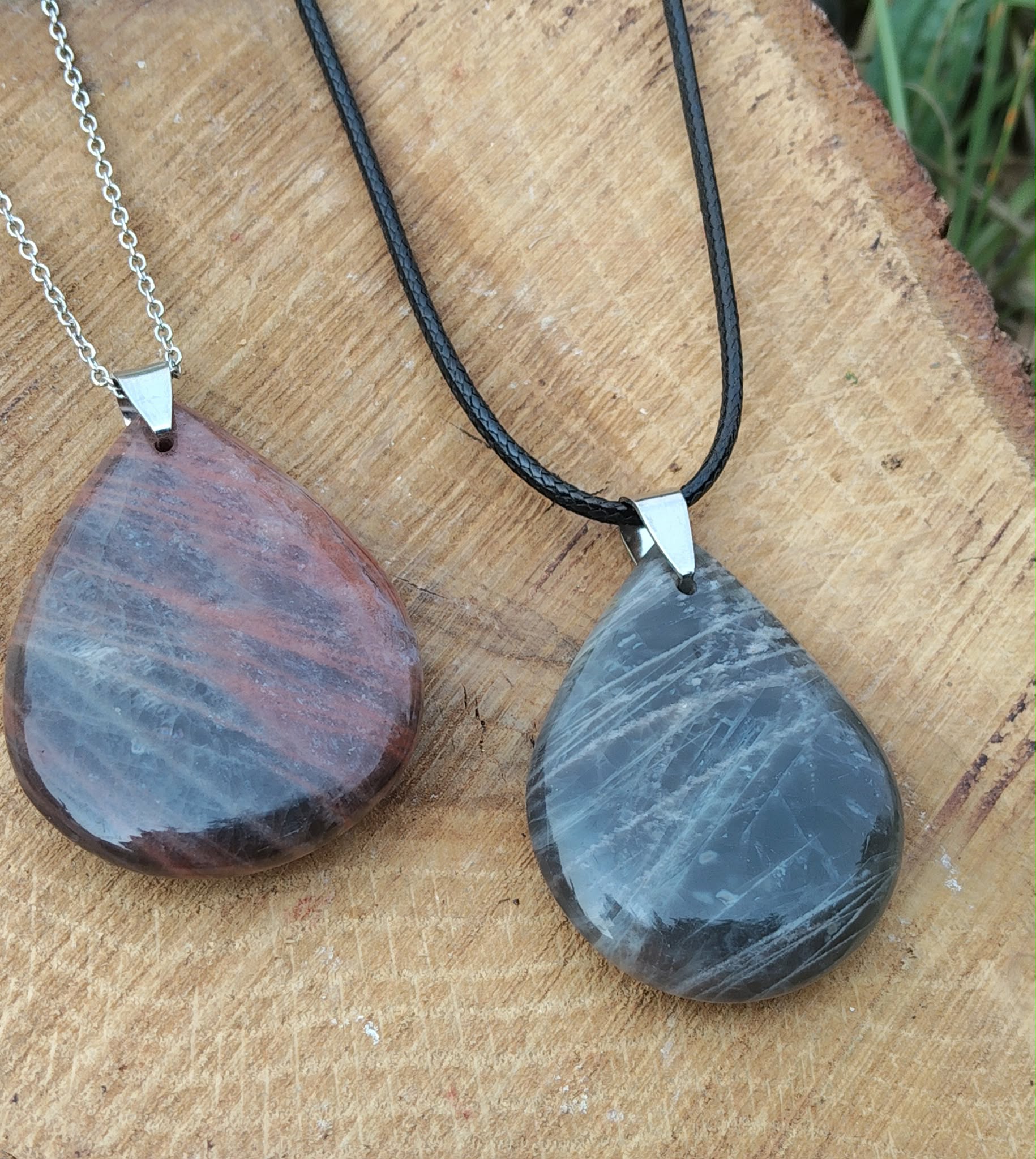Deux pendentifs goutte en pierre de lune noire, l’un sur chaîne argentée et l’autre sur cordon noir, posés sur une tranche de bois, bijoux en pierre naturelle associés au recentrage, à la douceur intérieure et à l’équilibre émotionnel.