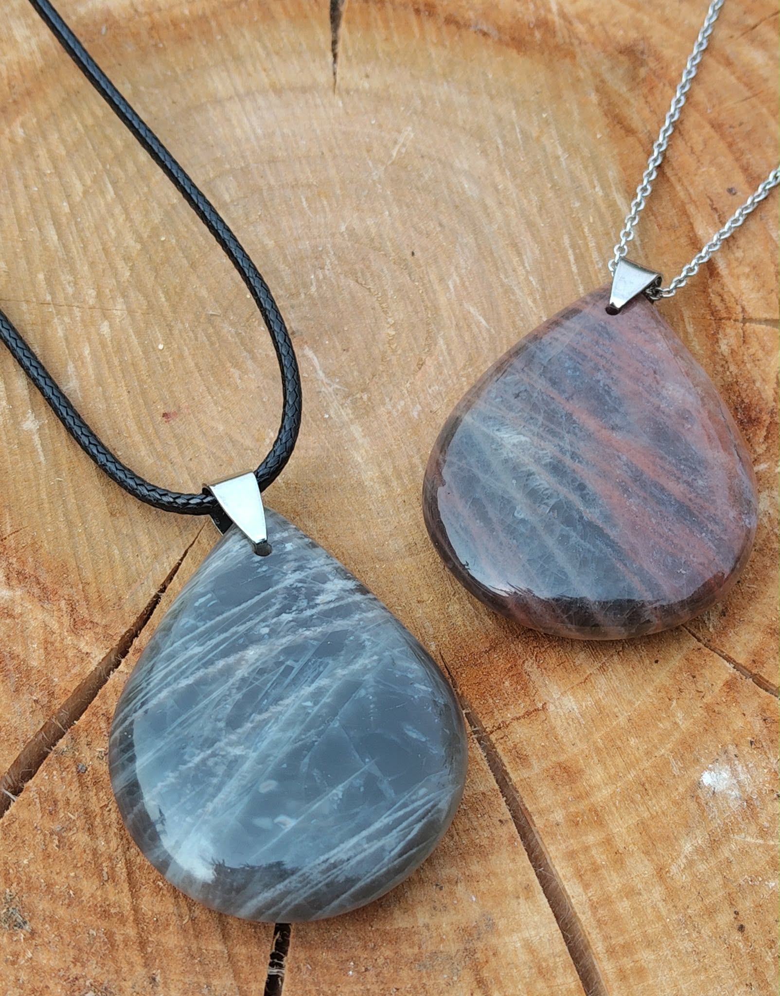 Deux pendentifs goutte en pierre de lune noire polie, l’un sur cordon noir et l’autre sur chaîne argentée, posés sur une tranche de bois, bijoux en pierre naturelle associés à la douceur émotionnelle, à l’ancrage et à l’harmonie intérieure.